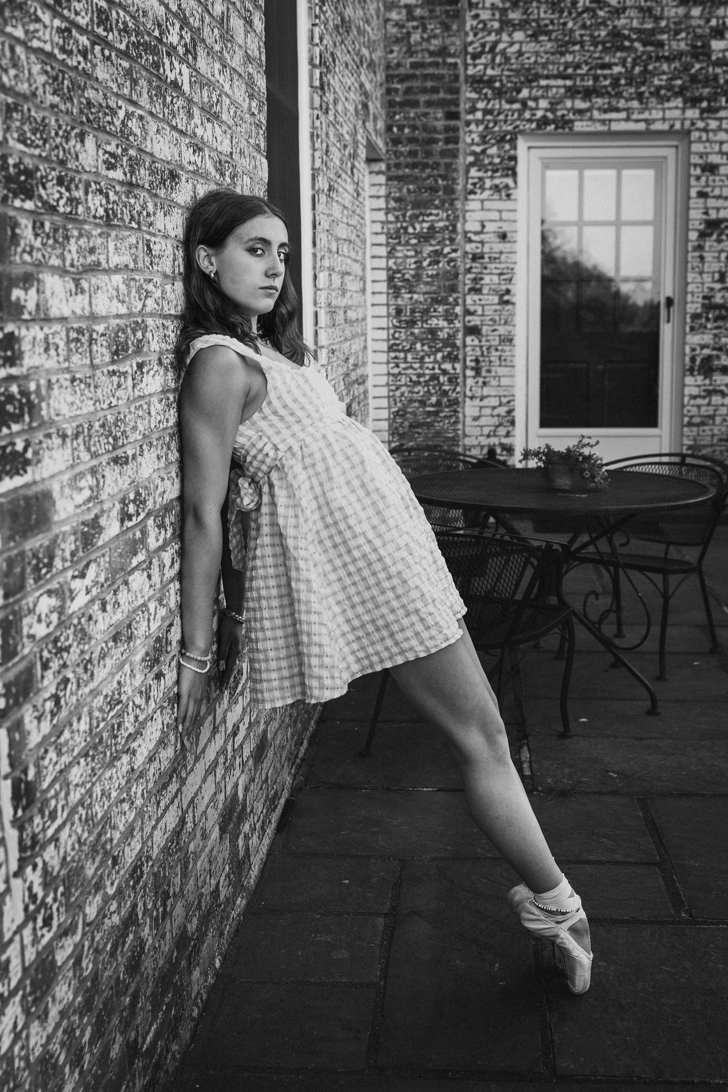 Black and white photo of a ballerina in a checkered dress posing on pointe shoes against a brick wall outdoors.