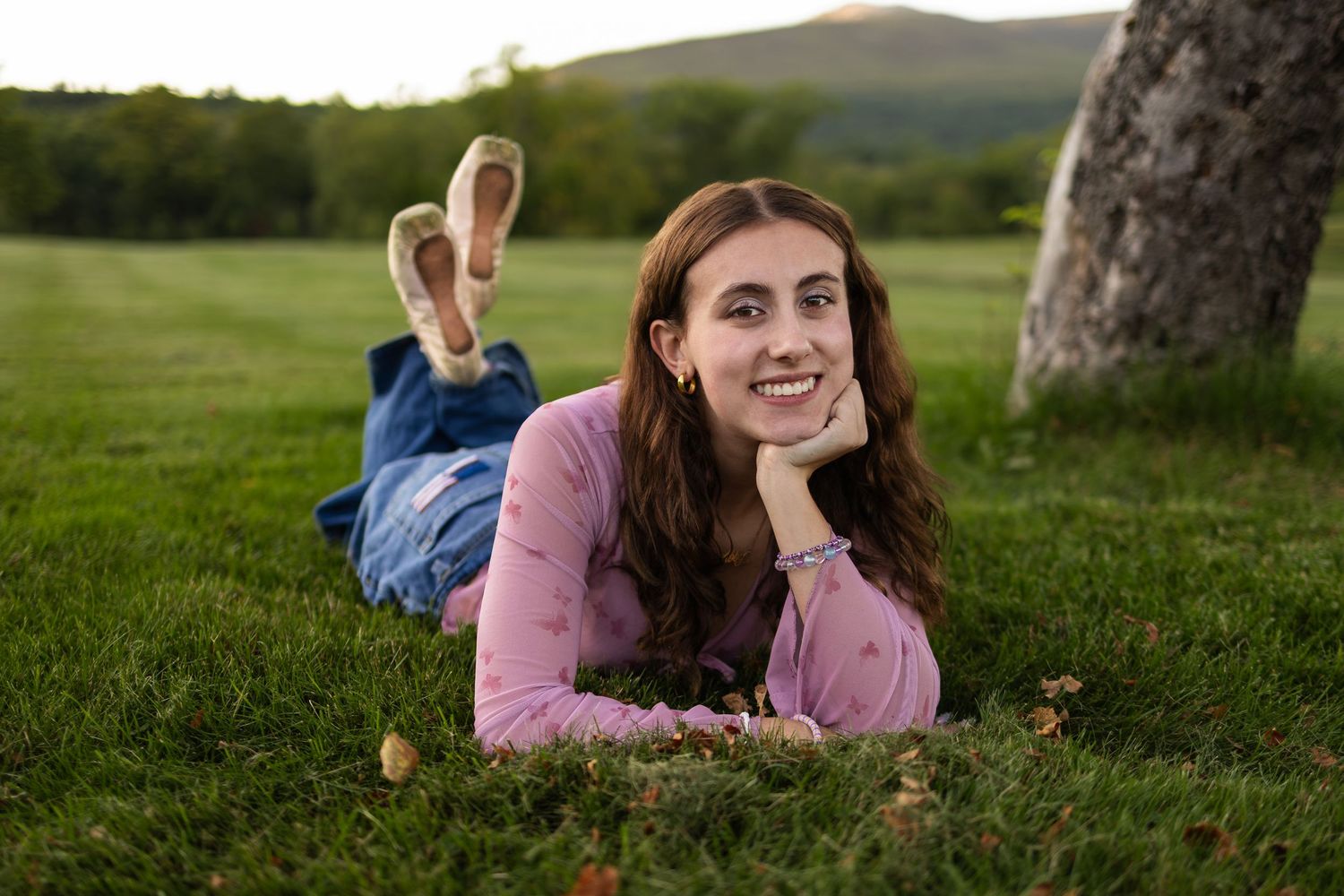 Young woman in pink top lying on green grass, smiling, with scenic mountain backdrop at sunset.