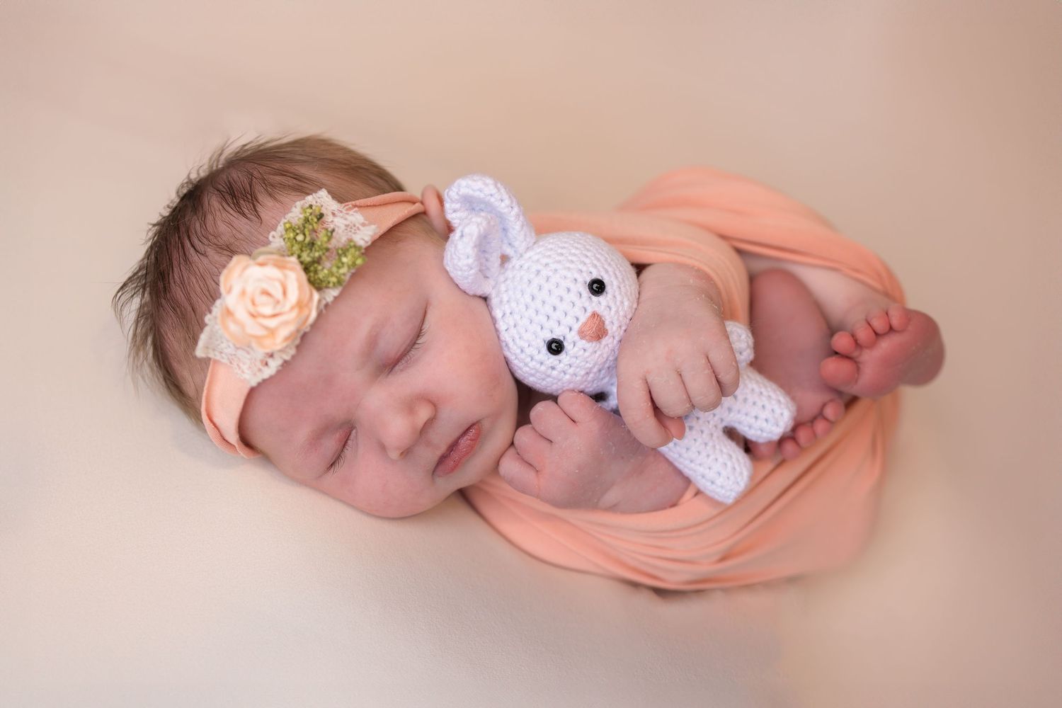 Newborn baby girl sleeping peacefully wrapped in peach swaddle, wearing floral headband, holding a crochet bunny toy.