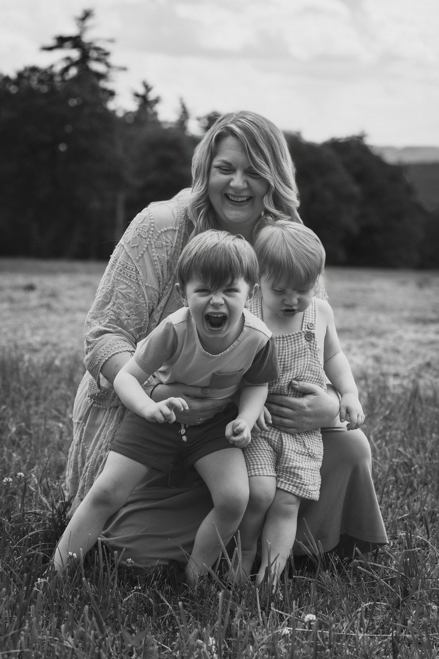 Black and white photo of a laughing mother kneeling in a field with her two young children outdoors.