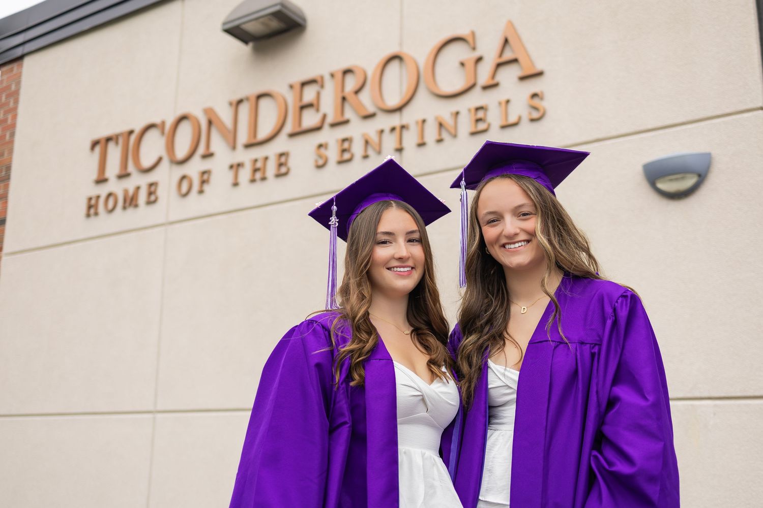 Two graduates in purple caps and gowns celebrate together in front of school building.