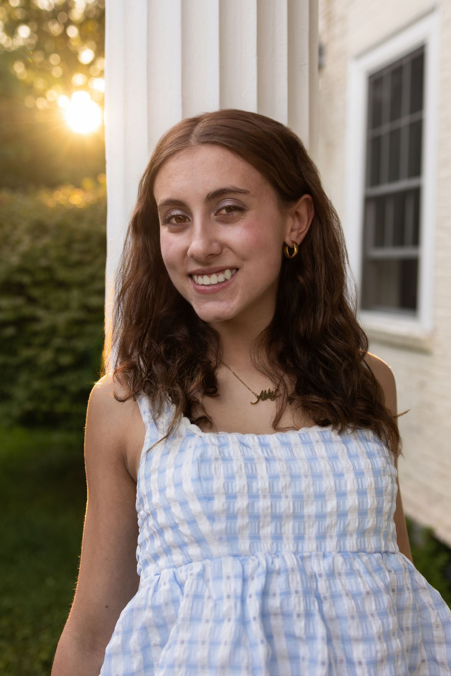 Young woman smiling in blue gingham dress, standing by white column at golden hour sunset.