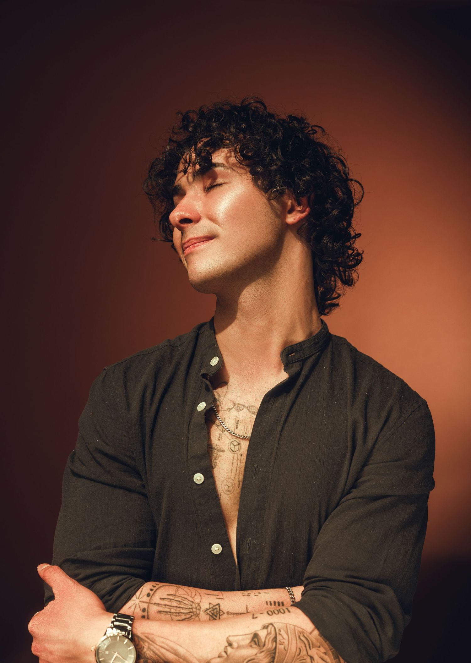 A person in a black button-up shirt with curly hair and tattoos poses against a dark reddish-brown background.
