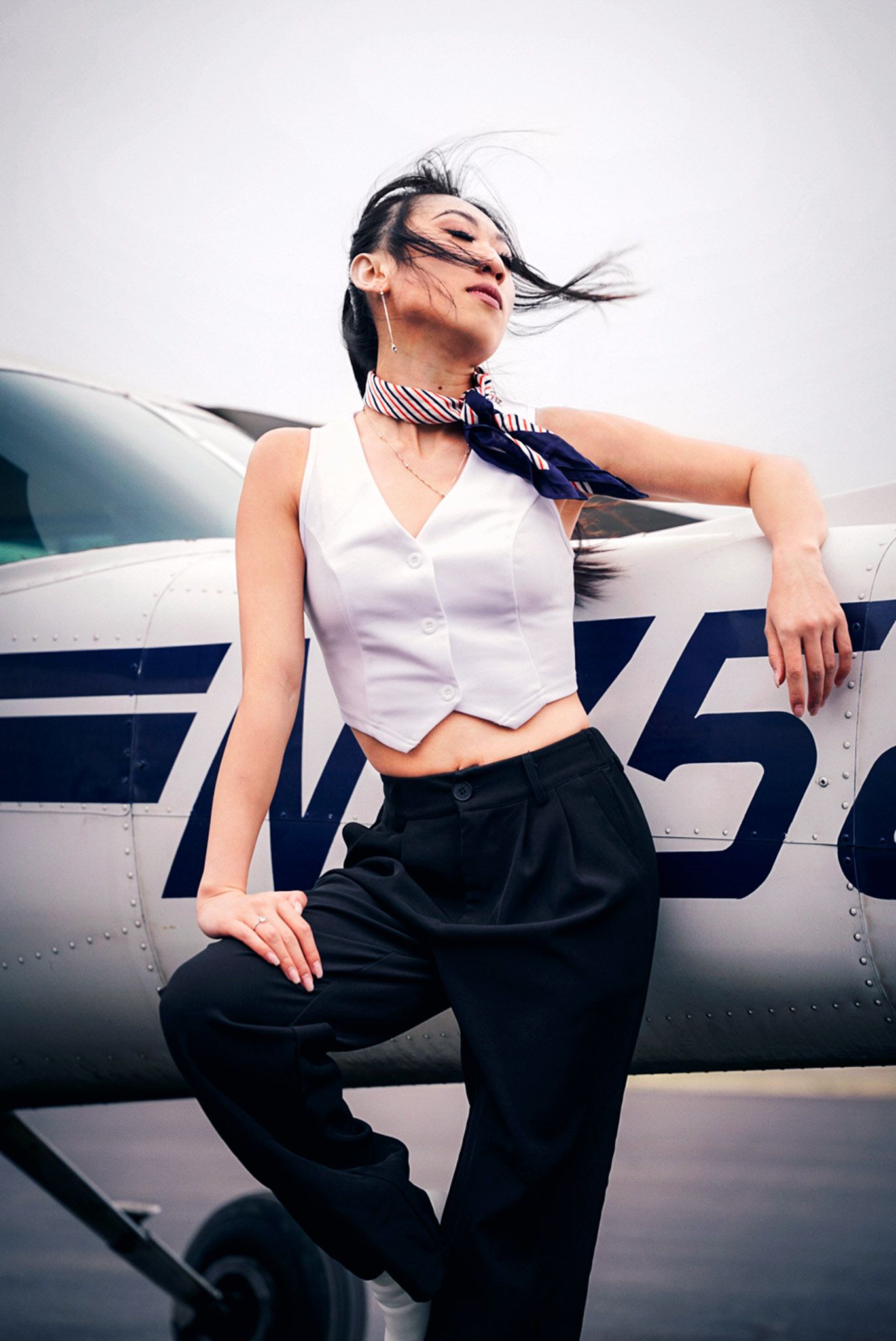 Fashion model in white crop top and black pants poses dramatically against small aircraft in moody photoshoot series.