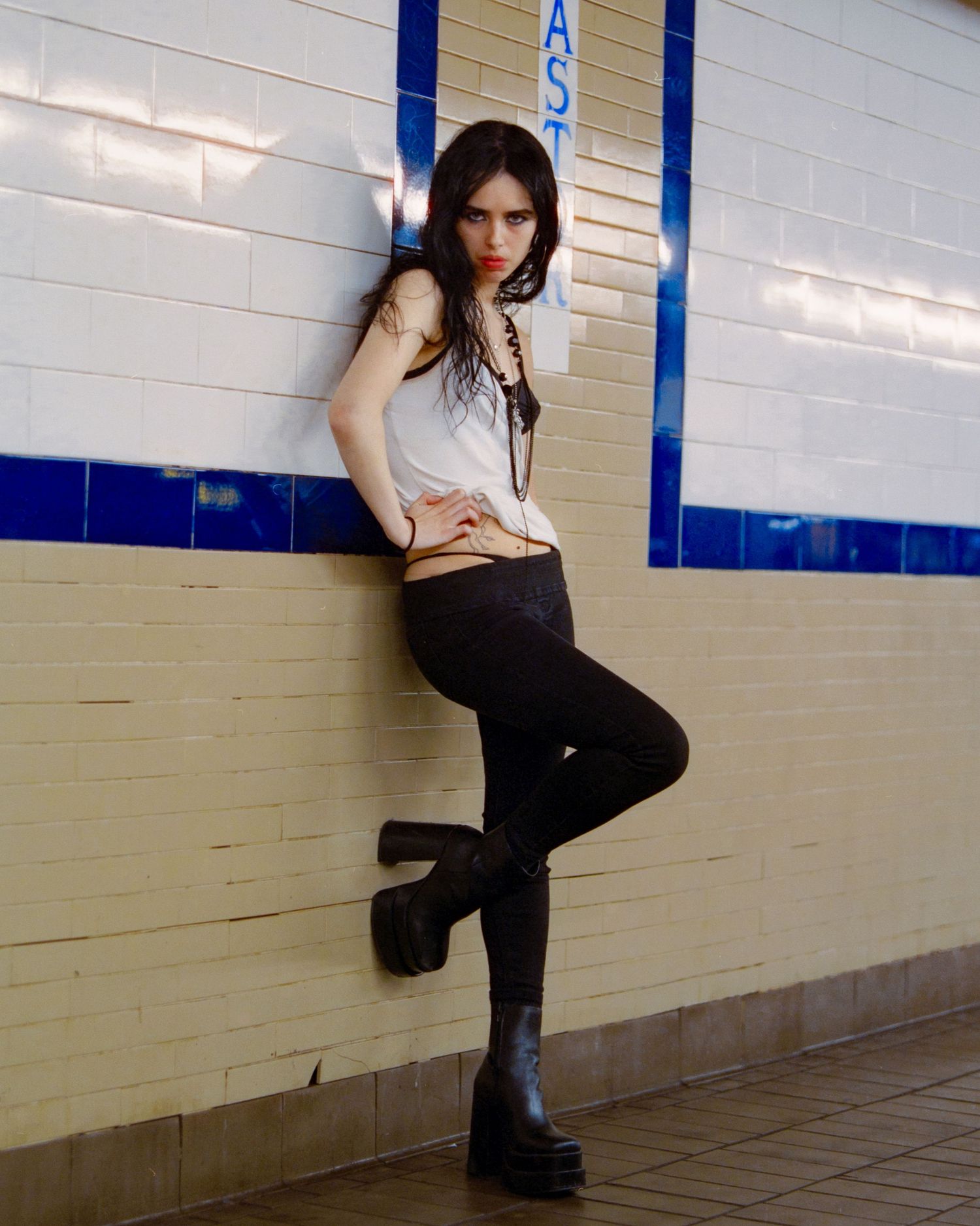 Young woman in black leggings and platform boots leans against tiled subway wall, striking a confident pose.