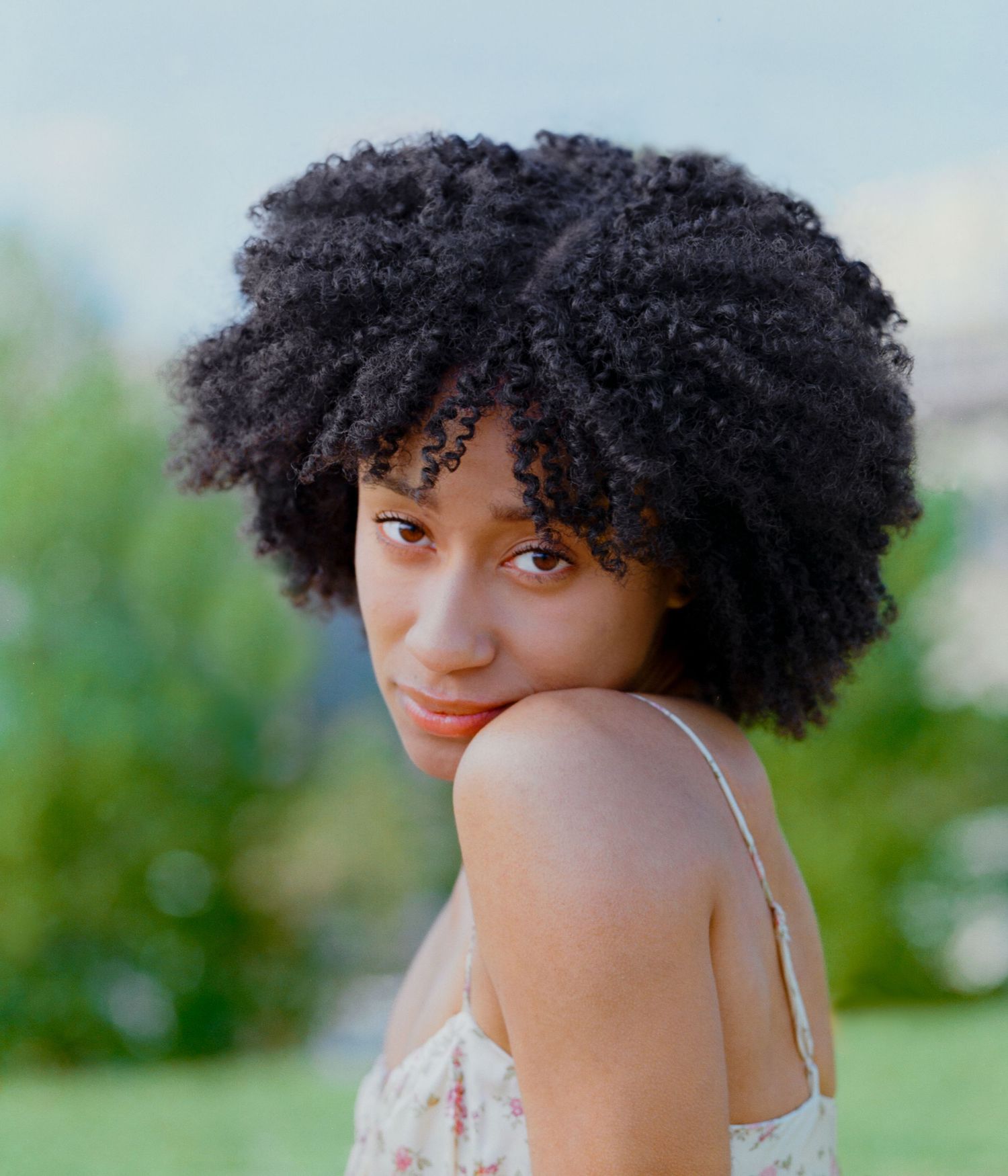 Young Black woman with natural afro hair wearing a floral dress, looking over her shoulder outdoors.