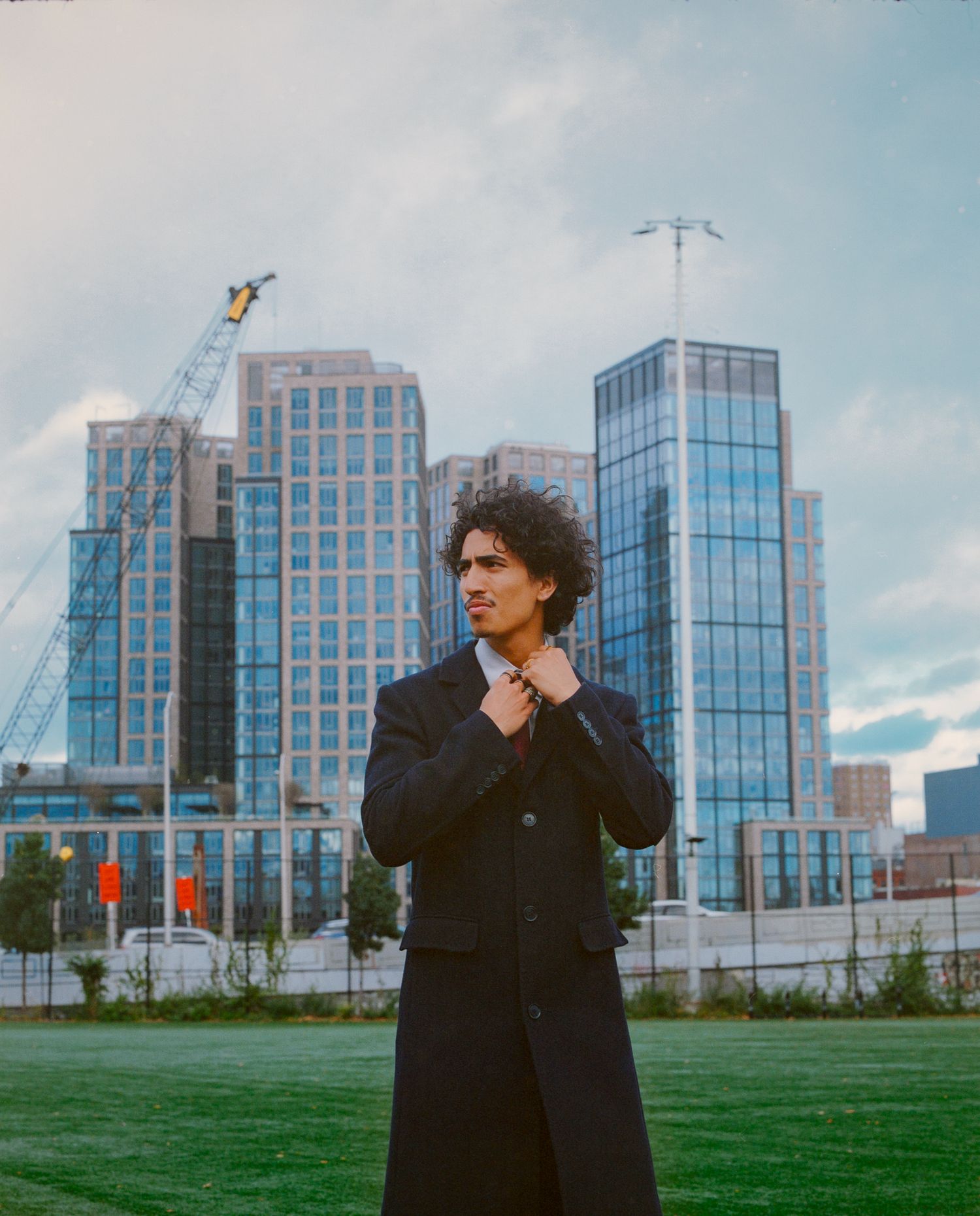 Figure in black coat stands against modern glass office buildings and green lawn.