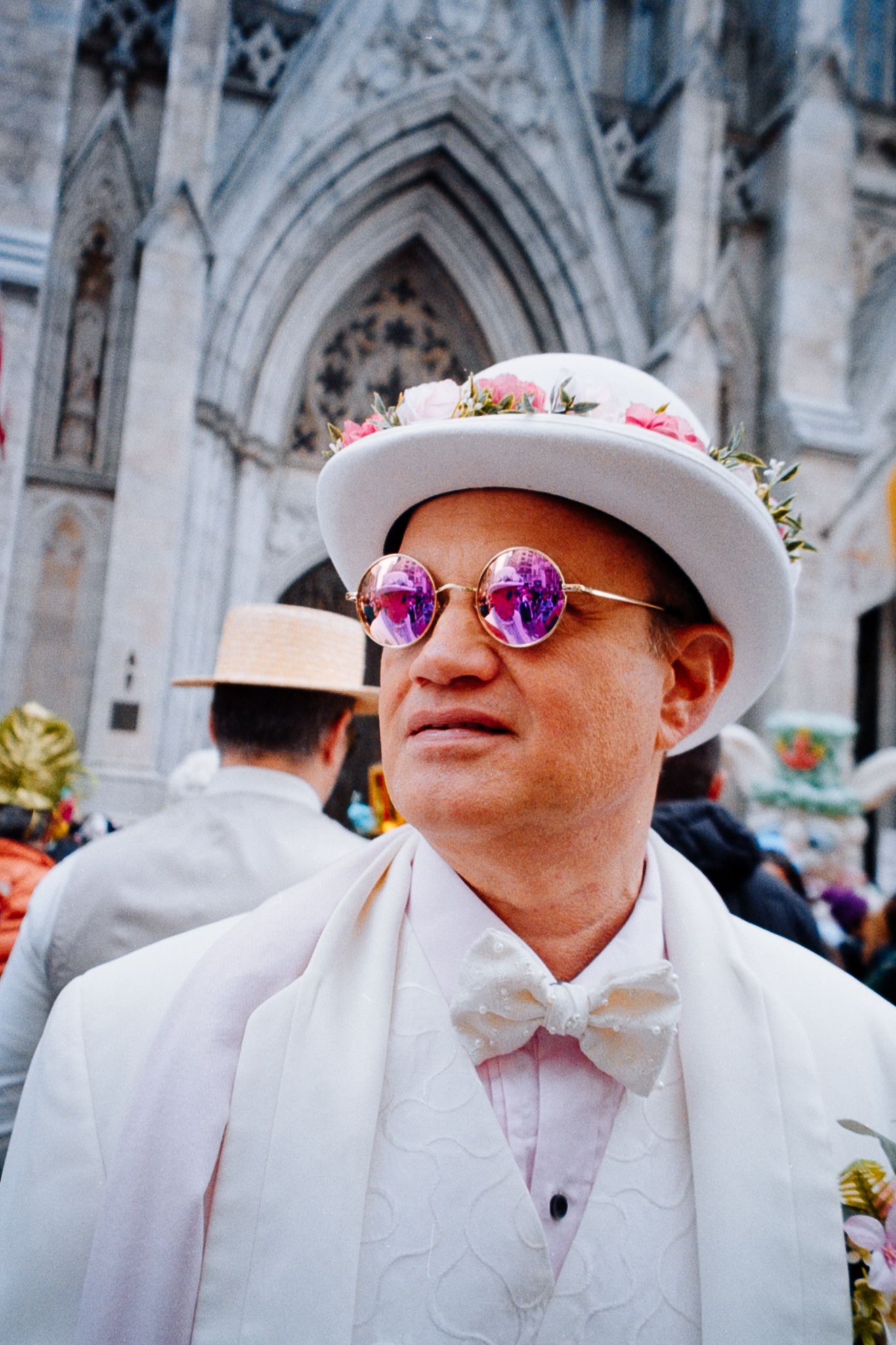 Man in white suit and floral hat with pink round sunglasses at Easter parade near cathedral.