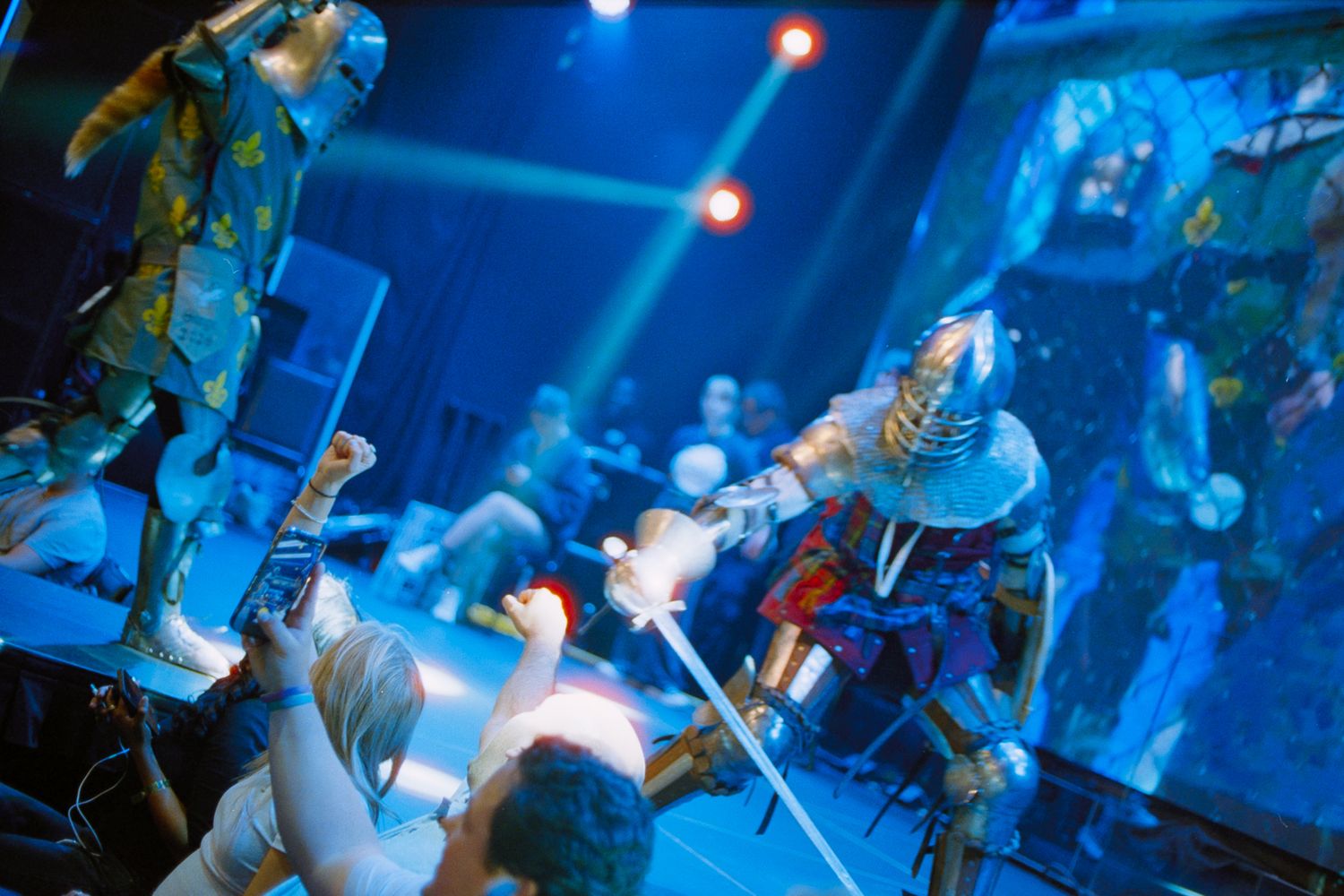 Costumed performers in elaborate armor costumes on stage under blue and red spotlights.