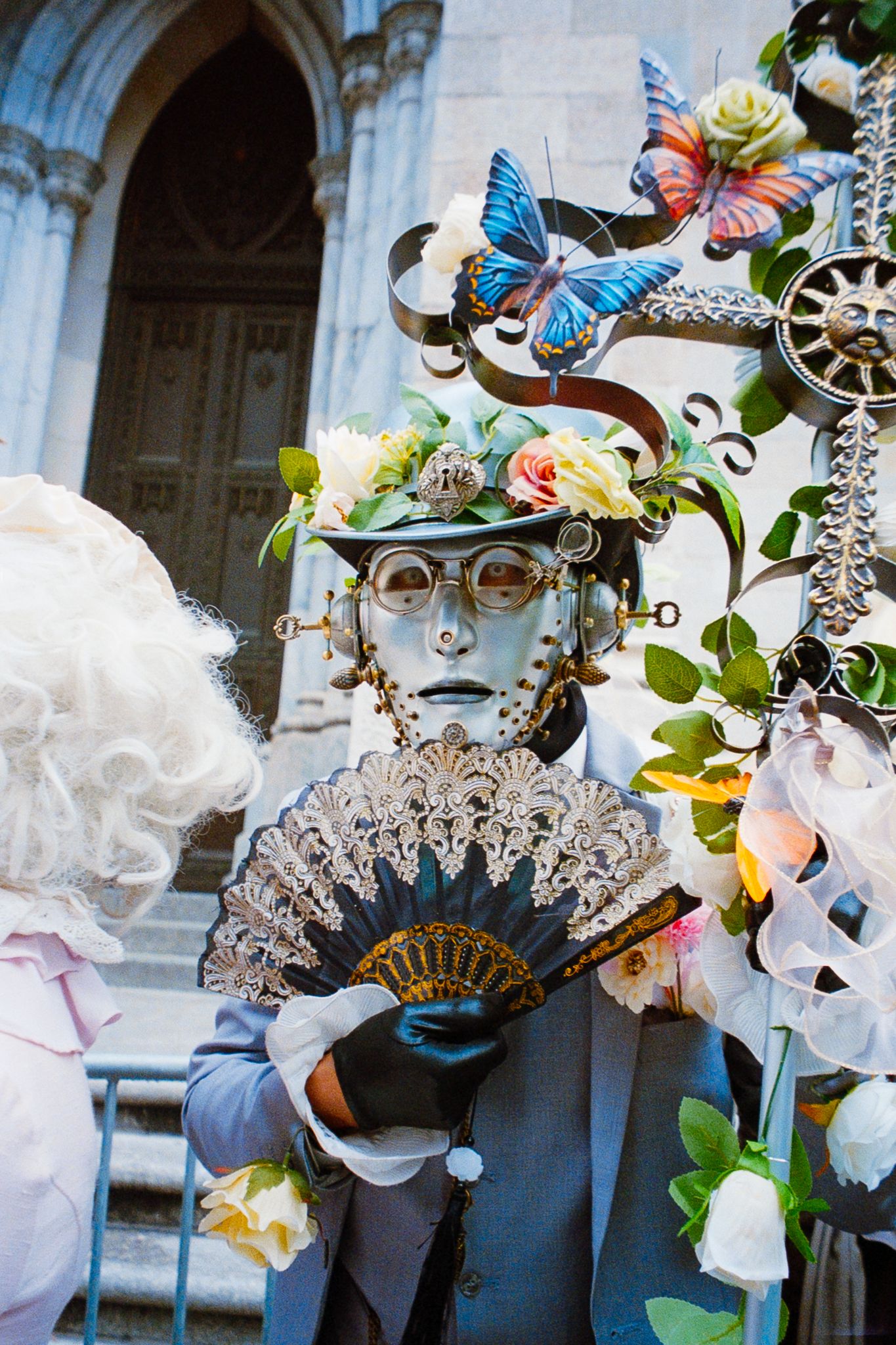 Elaborate steampunk costume with butterfly headpiece, goggles, and lace fan at a festive outdoor parade.
