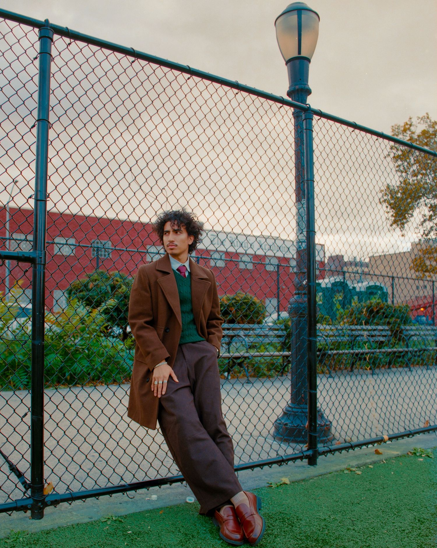 Person in brown coat leans against fence with urban waterfront view.