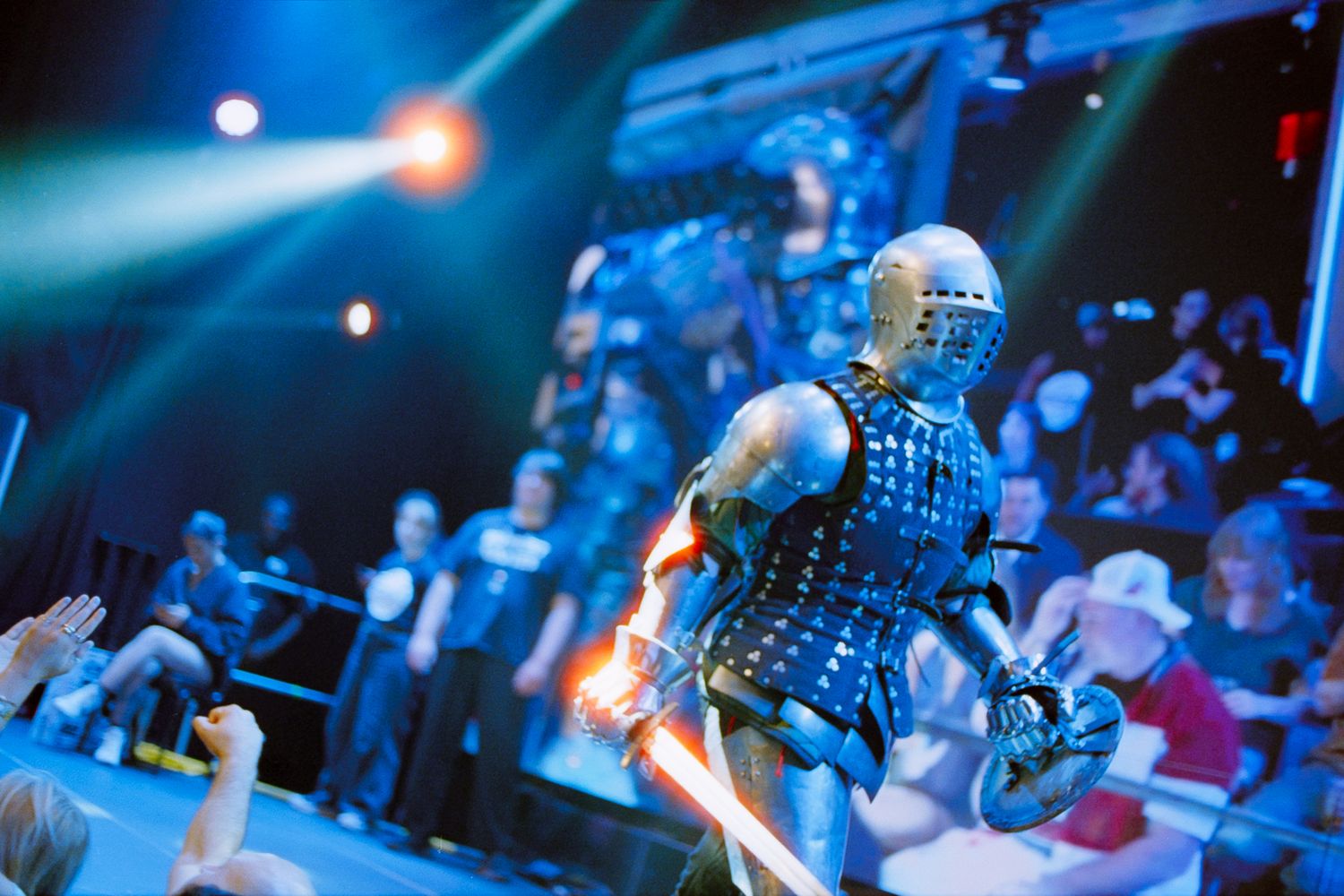 A medieval knight in armor performs on stage under blue spotlights before a live audience.