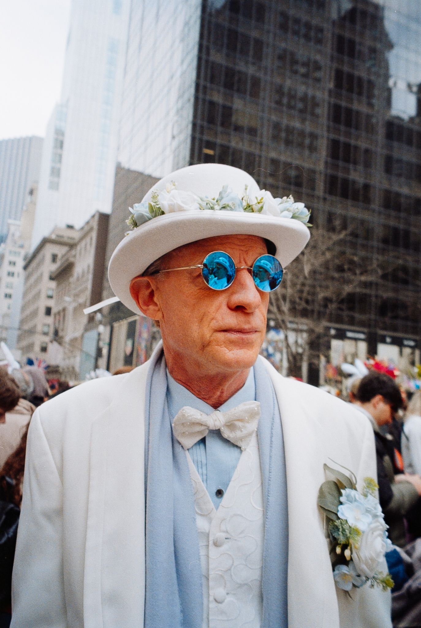 Elderly man in white suit and hat with blue round sunglasses at an outdoor Easter parade in New York City.