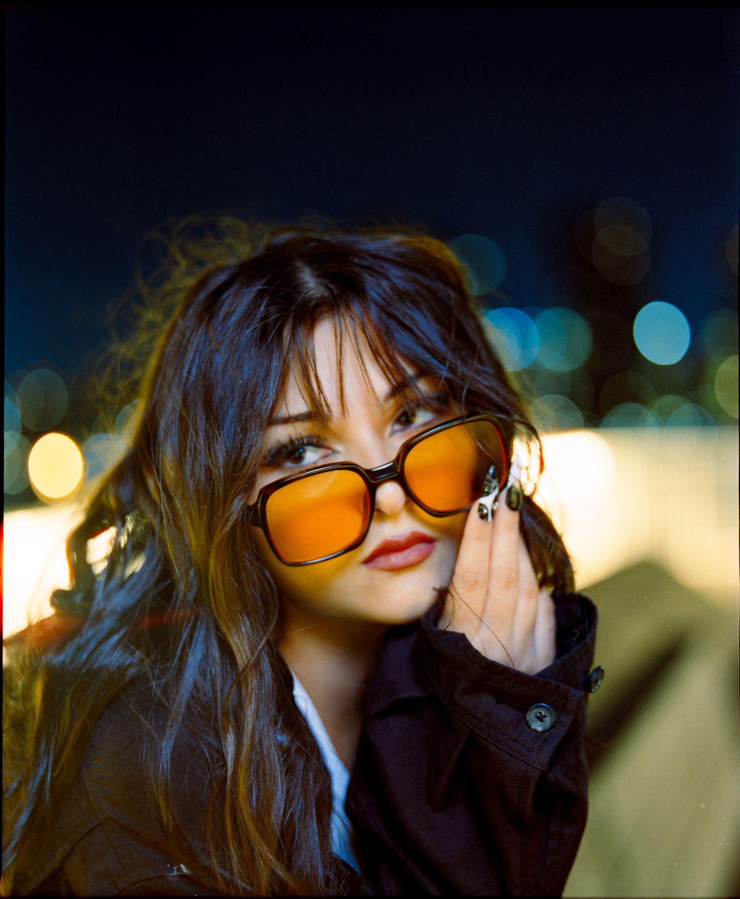 Stylish portrait with orange-tinted sunglasses and dark jacket in evening setting.