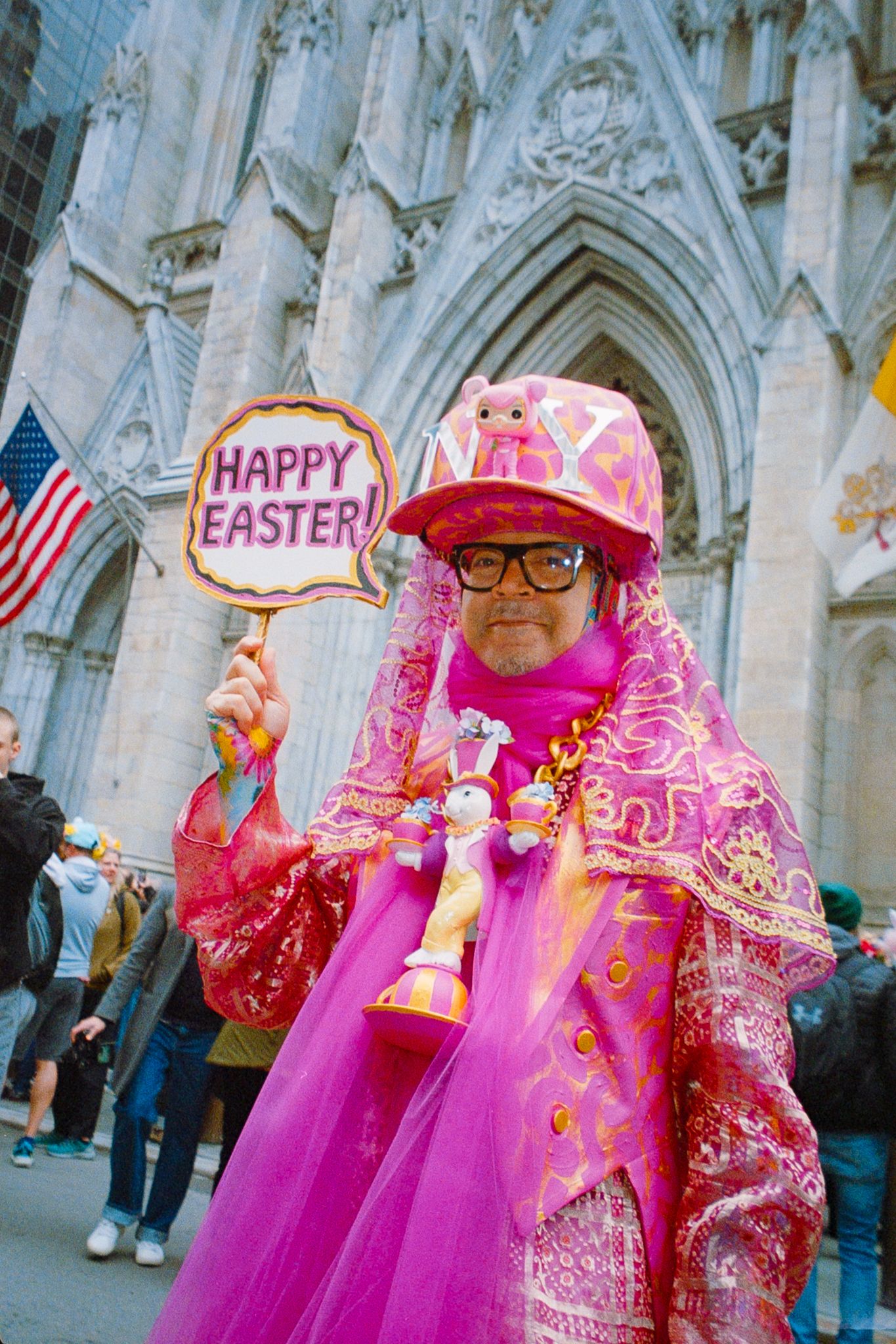 Person in vibrant pink Easter costume holding Happy Easter sign outside St. Patrick's Cathedral NYC.