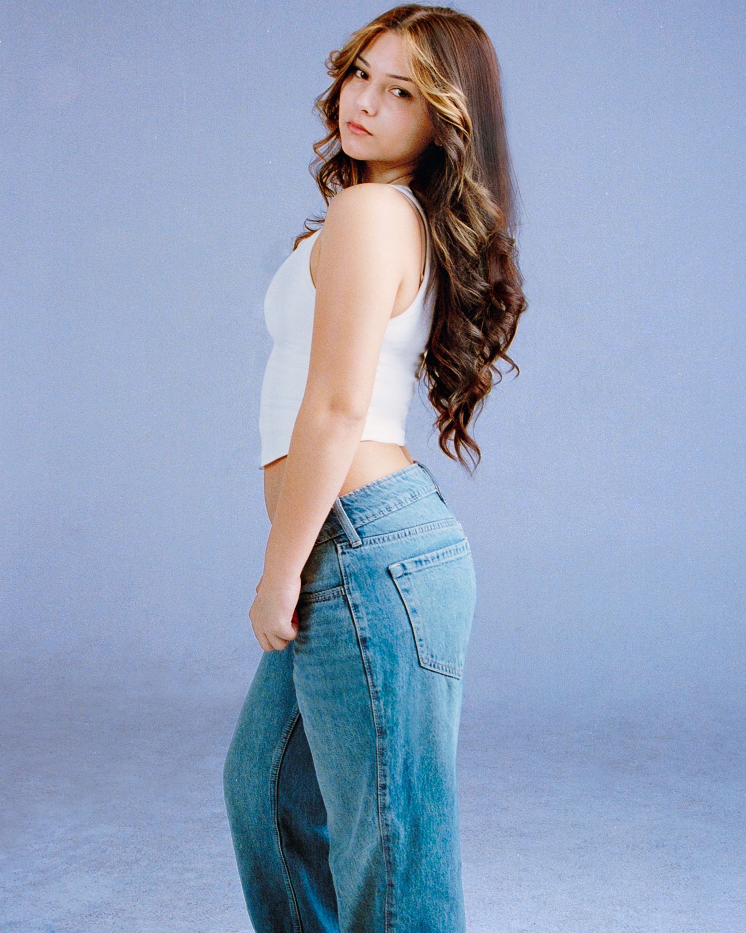 Young woman with long wavy brown hair wearing a white crop top and blue jeans posing against a gray background.