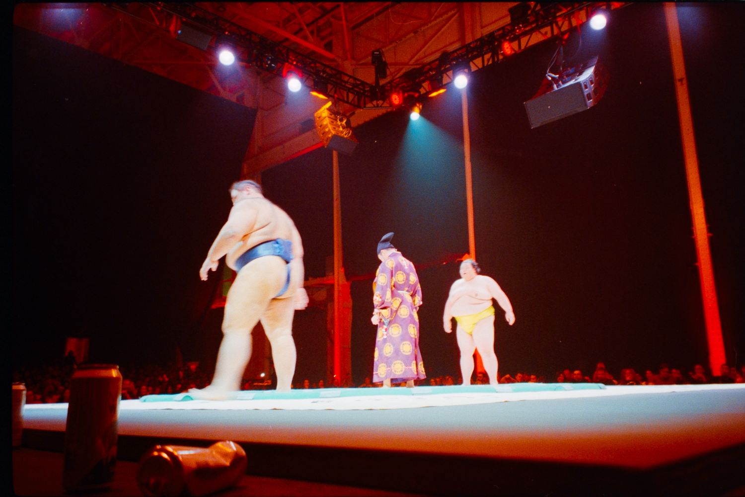 Three performers on a lit stage, two in sumo wrestler attire and one in a colorful robe, during a live show.