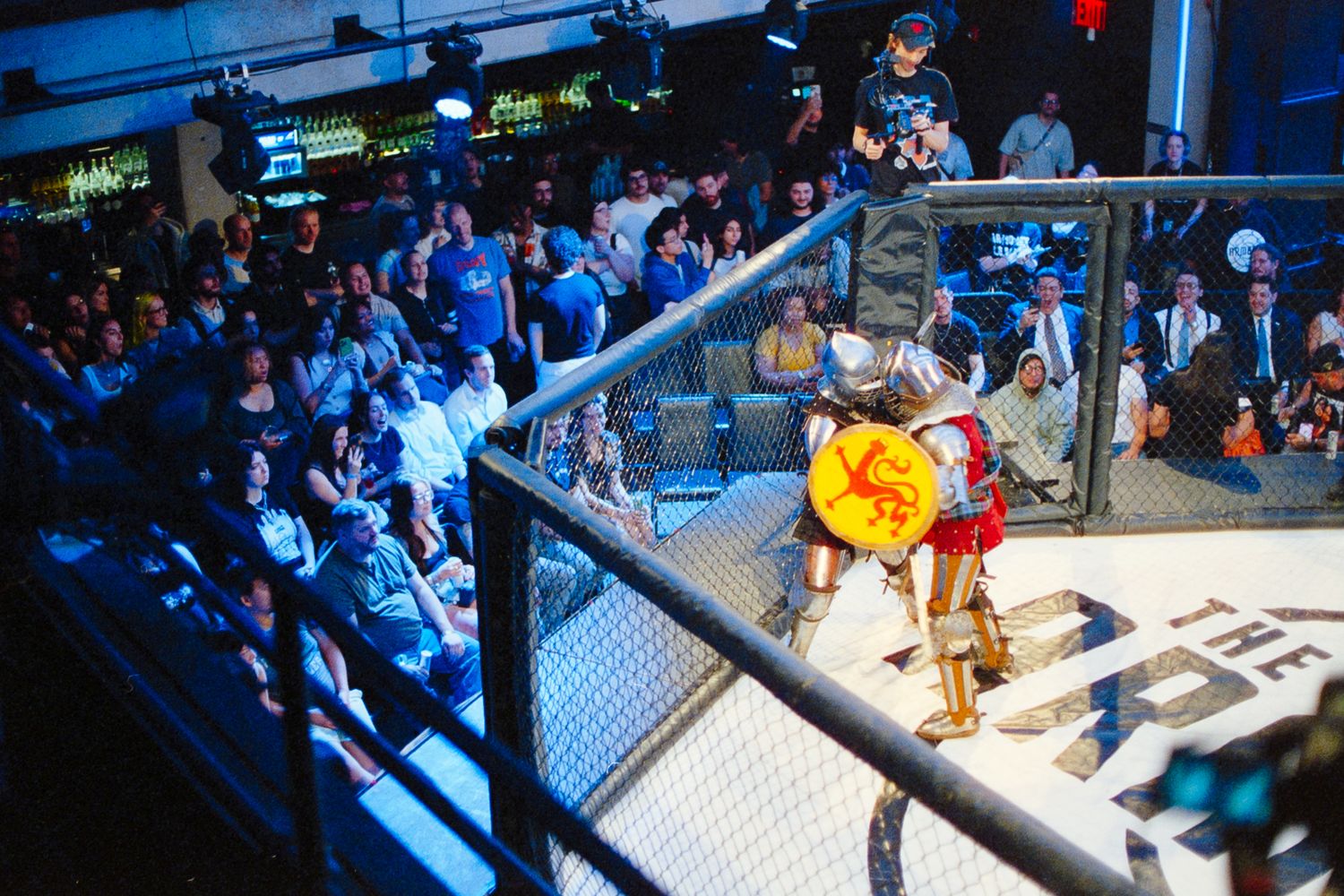Two fighters in medieval armor battle inside an MMA cage ring before a packed crowd at a live event.