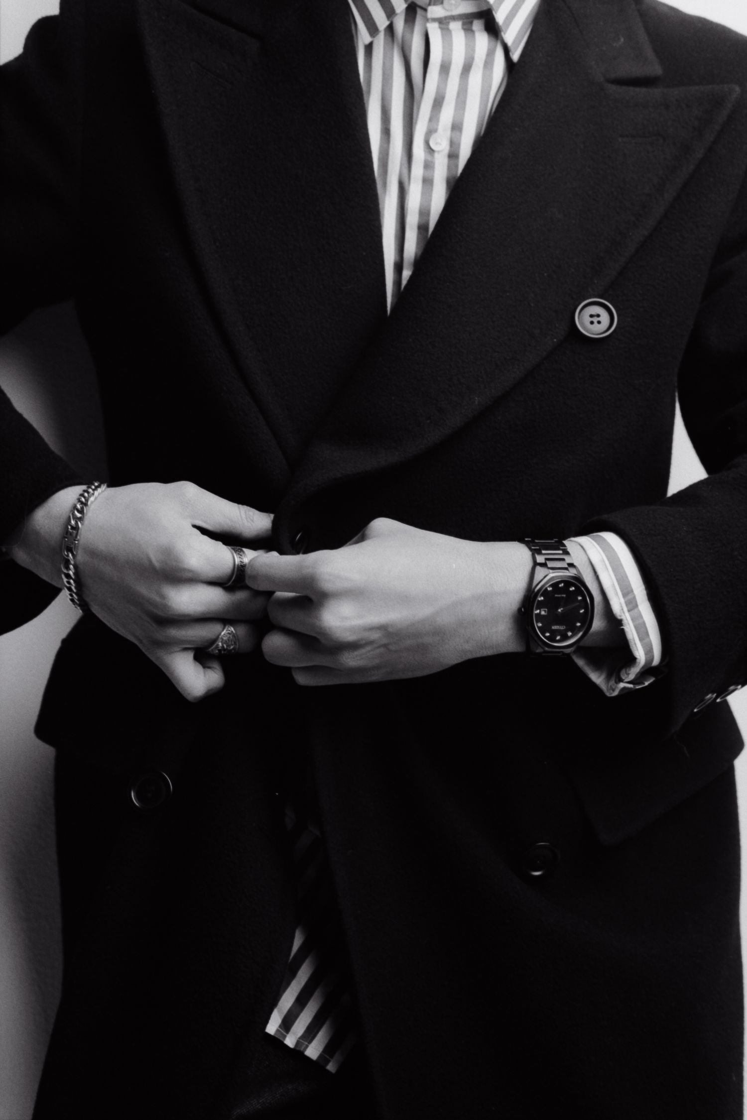 Black and white photo of a man in a double-breasted blazer buttoning his jacket, wearing a watch and bracelet.