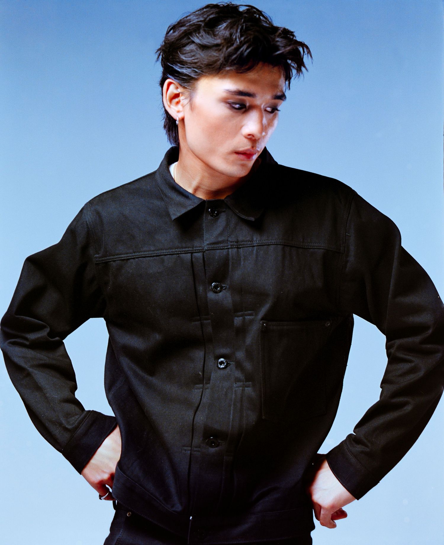 Young person with dark hair wearing a black button-up jacket, posing with hands on hips against a blue background.