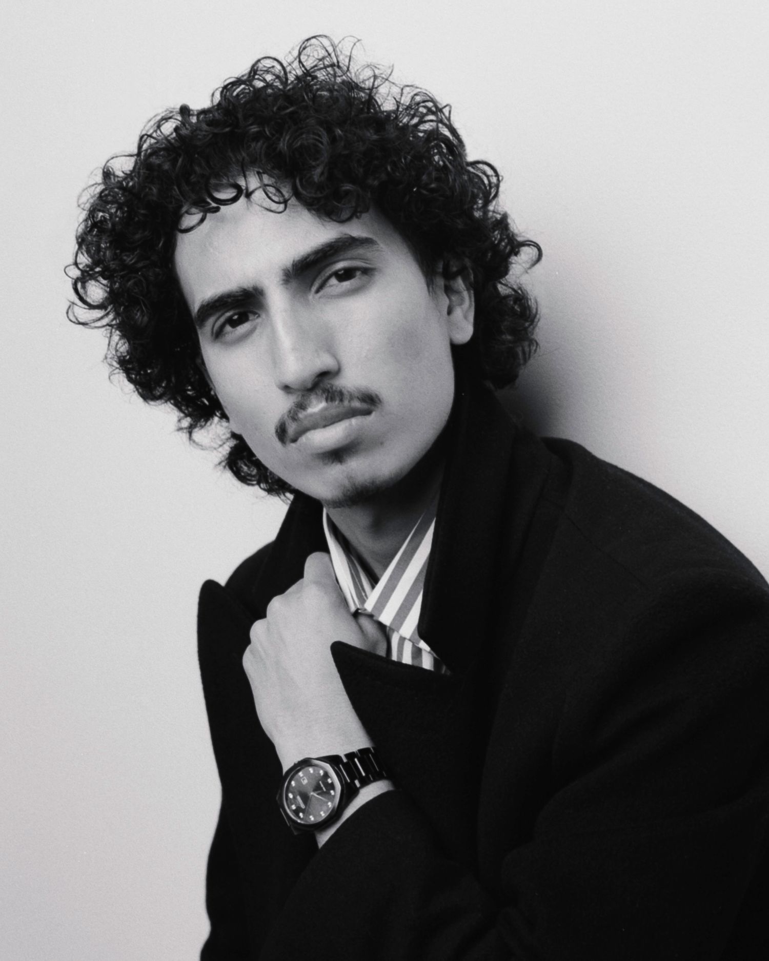 Black and white portrait of a young man with curly hair wearing a black coat and watch, posing confidently.