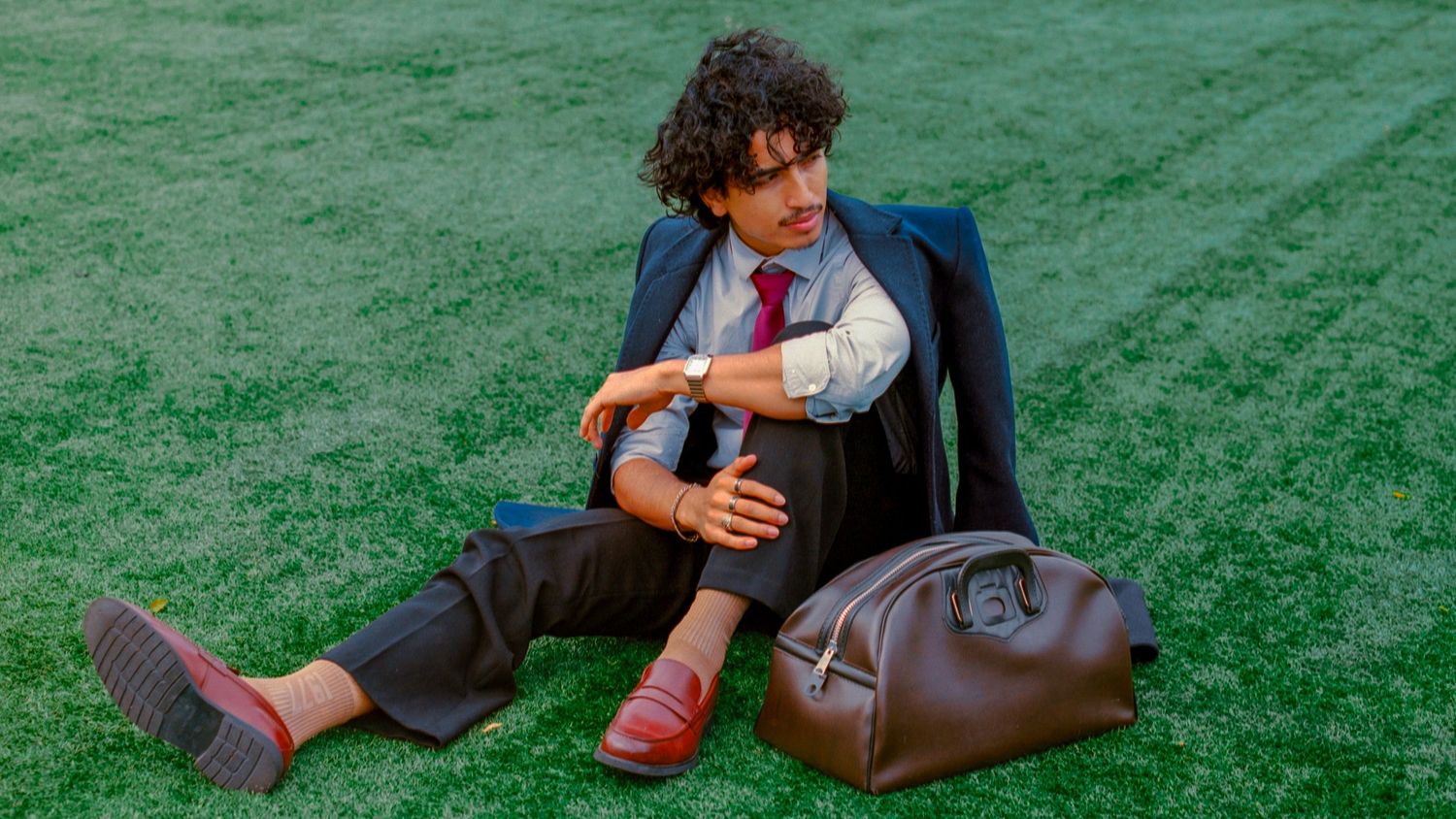 Young man in suit and red loafers sitting on green grass beside a brown leather duffle bag.