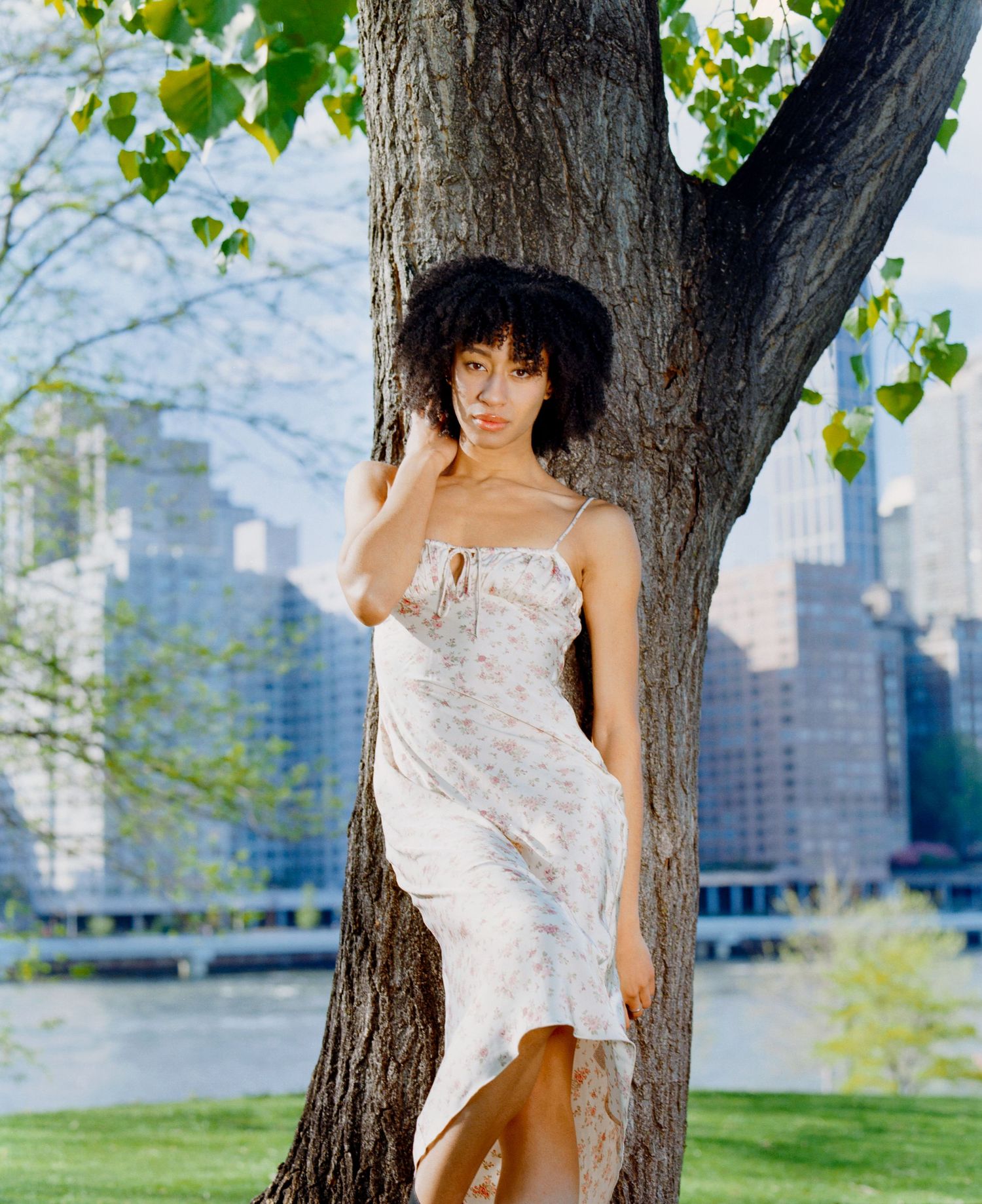 Woman in floral slip dress leaning against tree in urban park with city skyline in background.