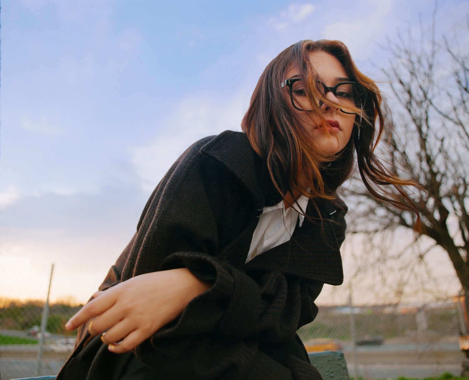 Young woman with glasses and windswept brown hair poses outdoors in a black coat at sunset.