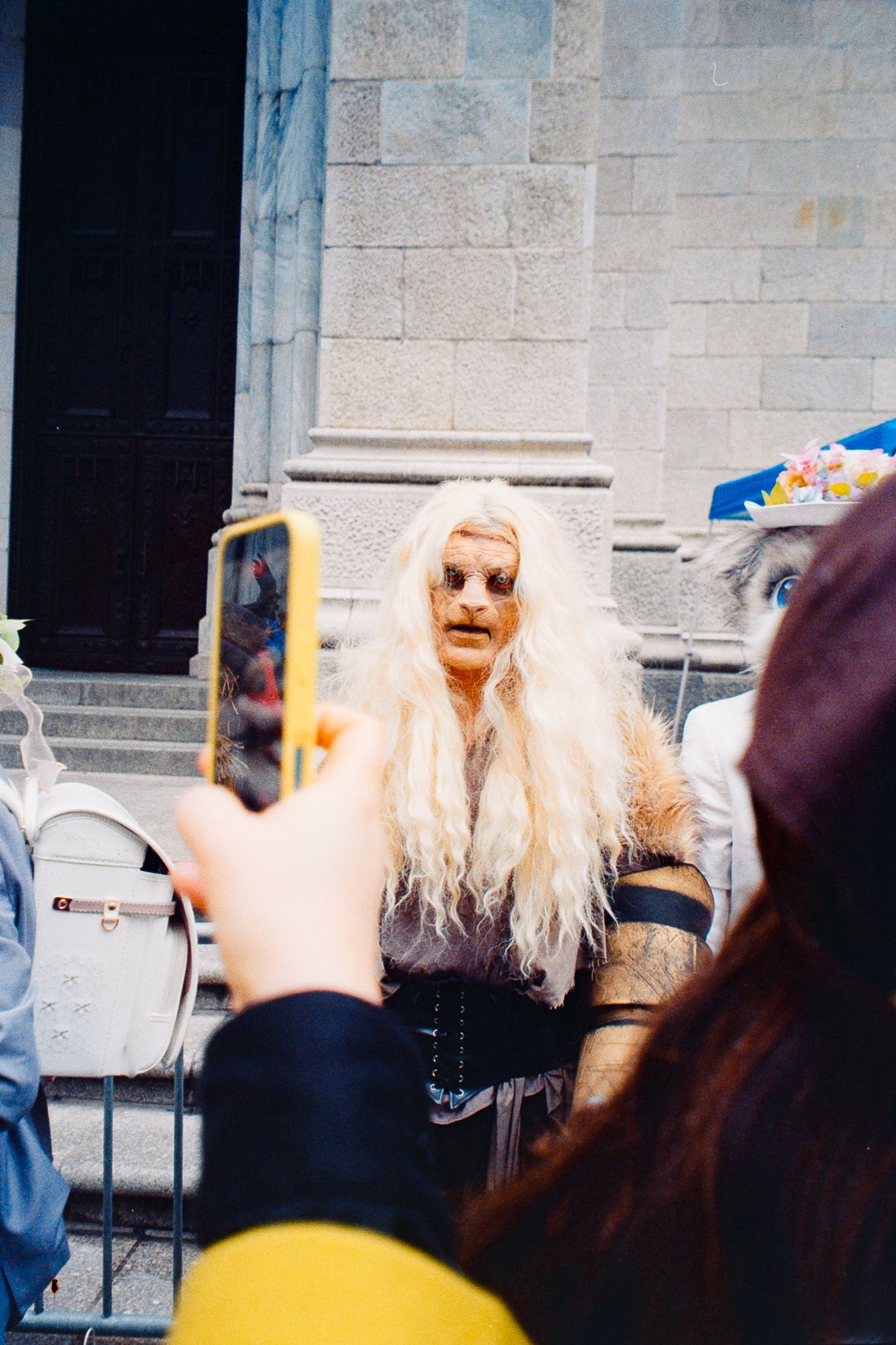 Person in dramatic white wig and dark costume being photographed on a smartphone at an outdoor event.