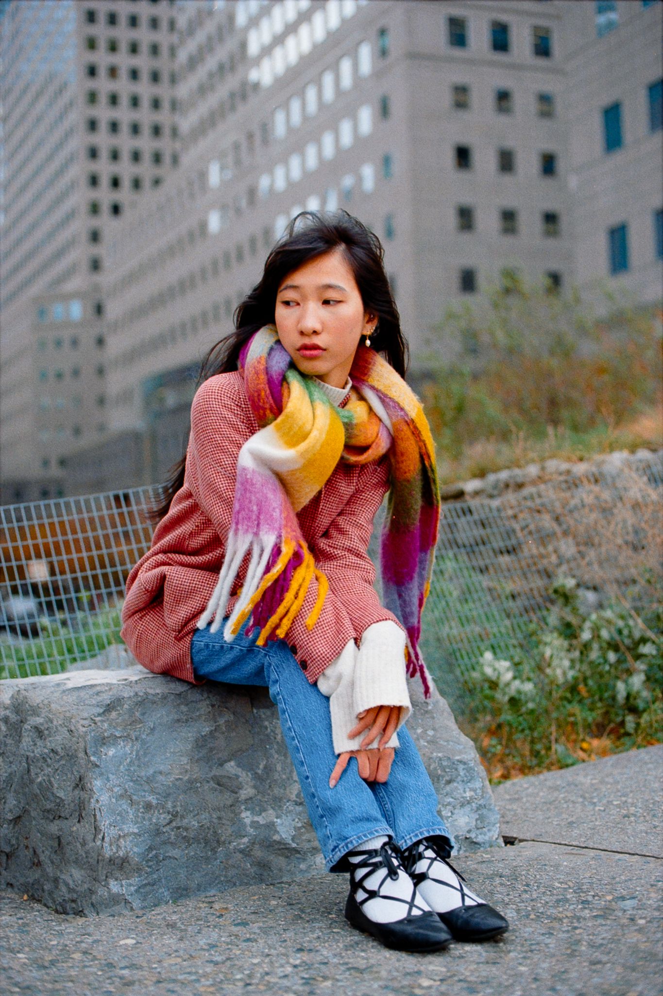 Model in pink coat and jeans poses casually on concrete ledge with city buildings in background.