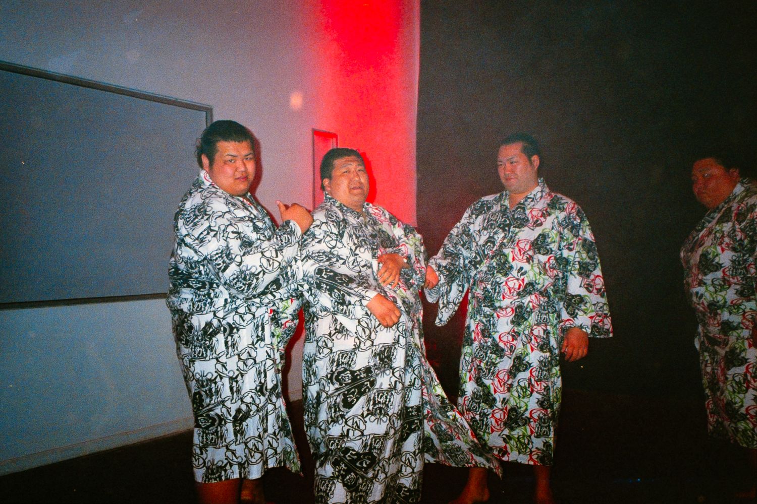 Four people wearing colorful floral robes stand together in a dimly lit room with red ambient lighting.