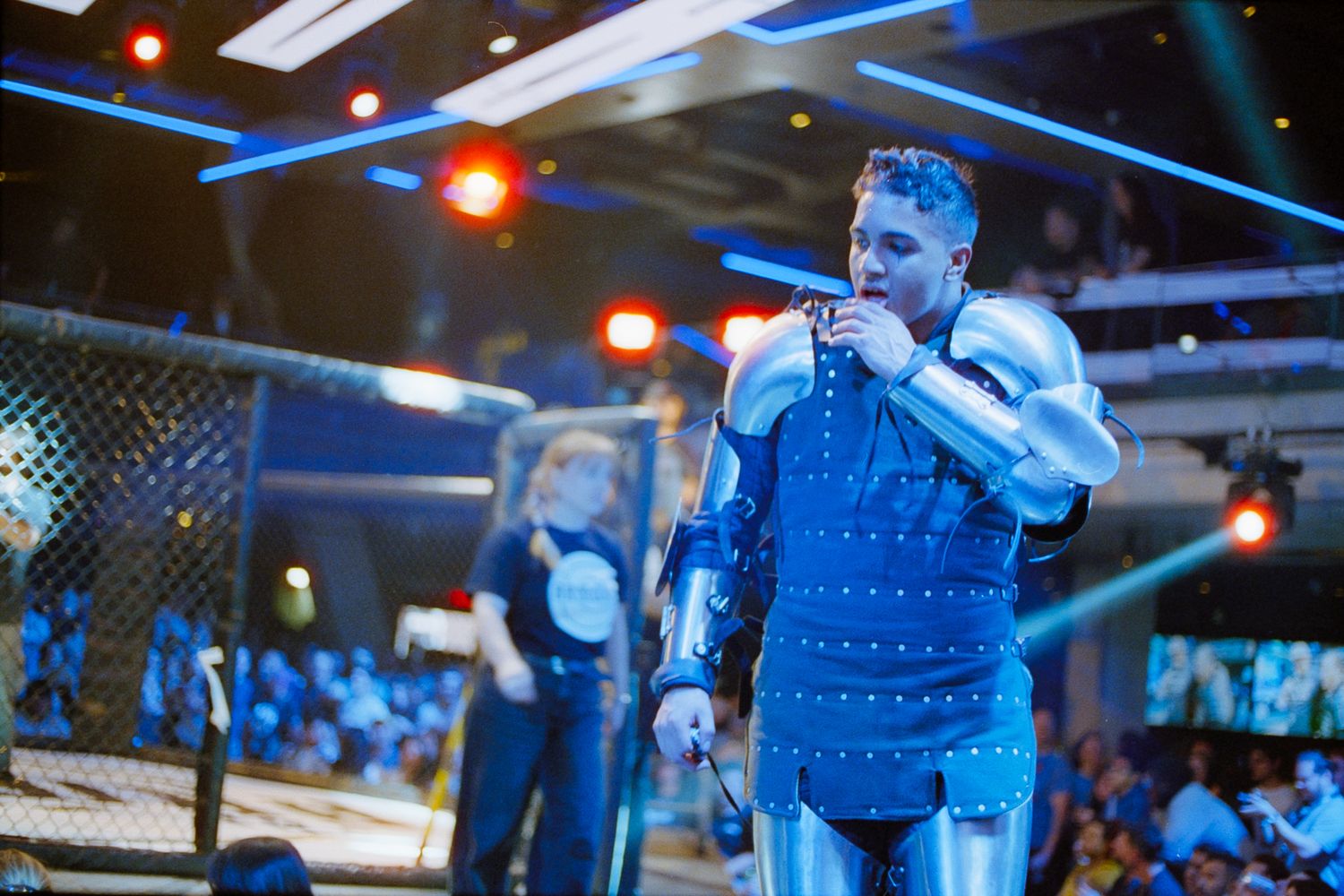 A performer in silver knight armor stands confidently on stage under blue lighting at a live event.