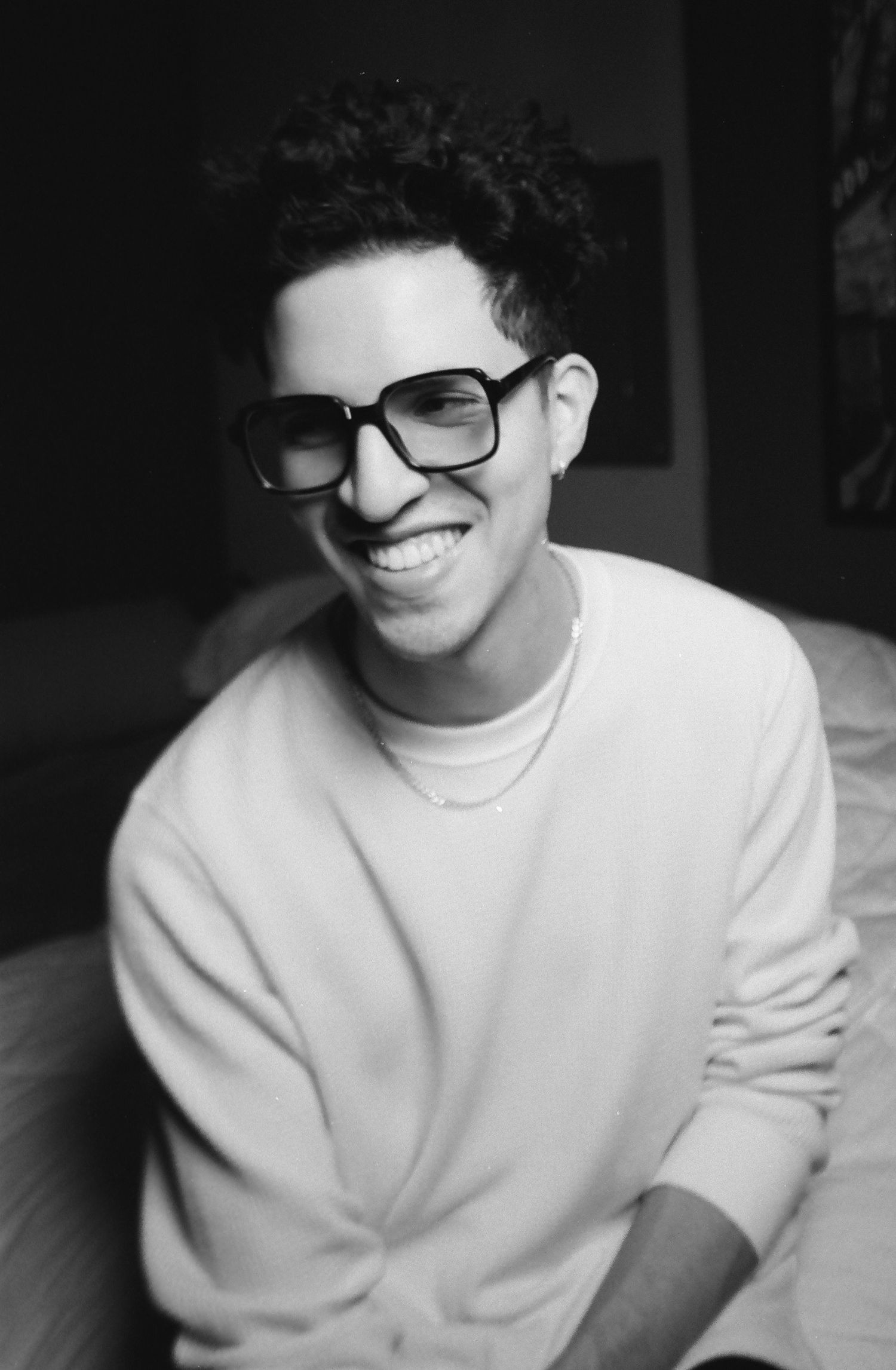 Smiling young man with curly hair wearing black glasses and a white sweatshirt in a black and white photo.