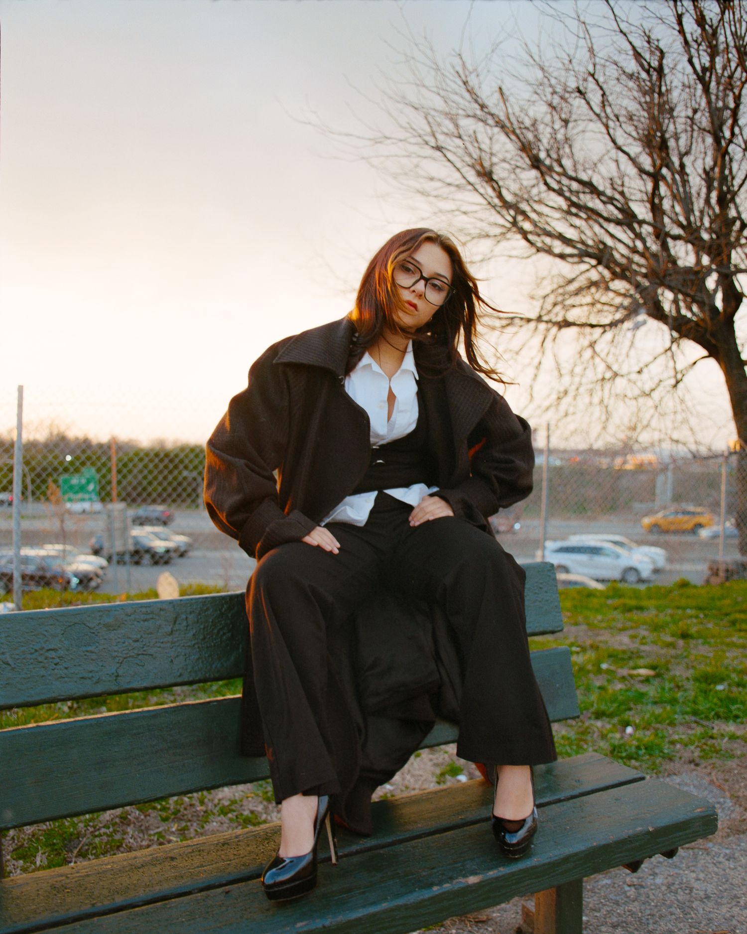 Woman in black oversized jacket and wide-leg pants sitting on a park bench at sunset wearing glasses.