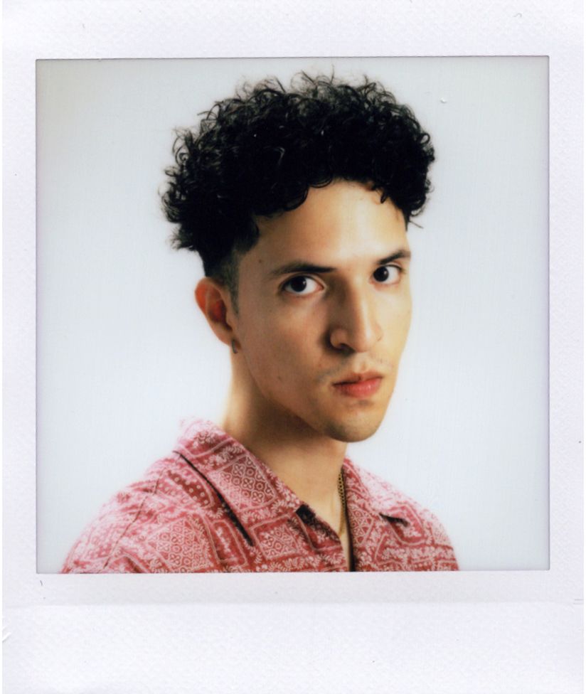 Young man with curly dark hair wearing a pink textured shirt, photographed in Polaroid style portrait.