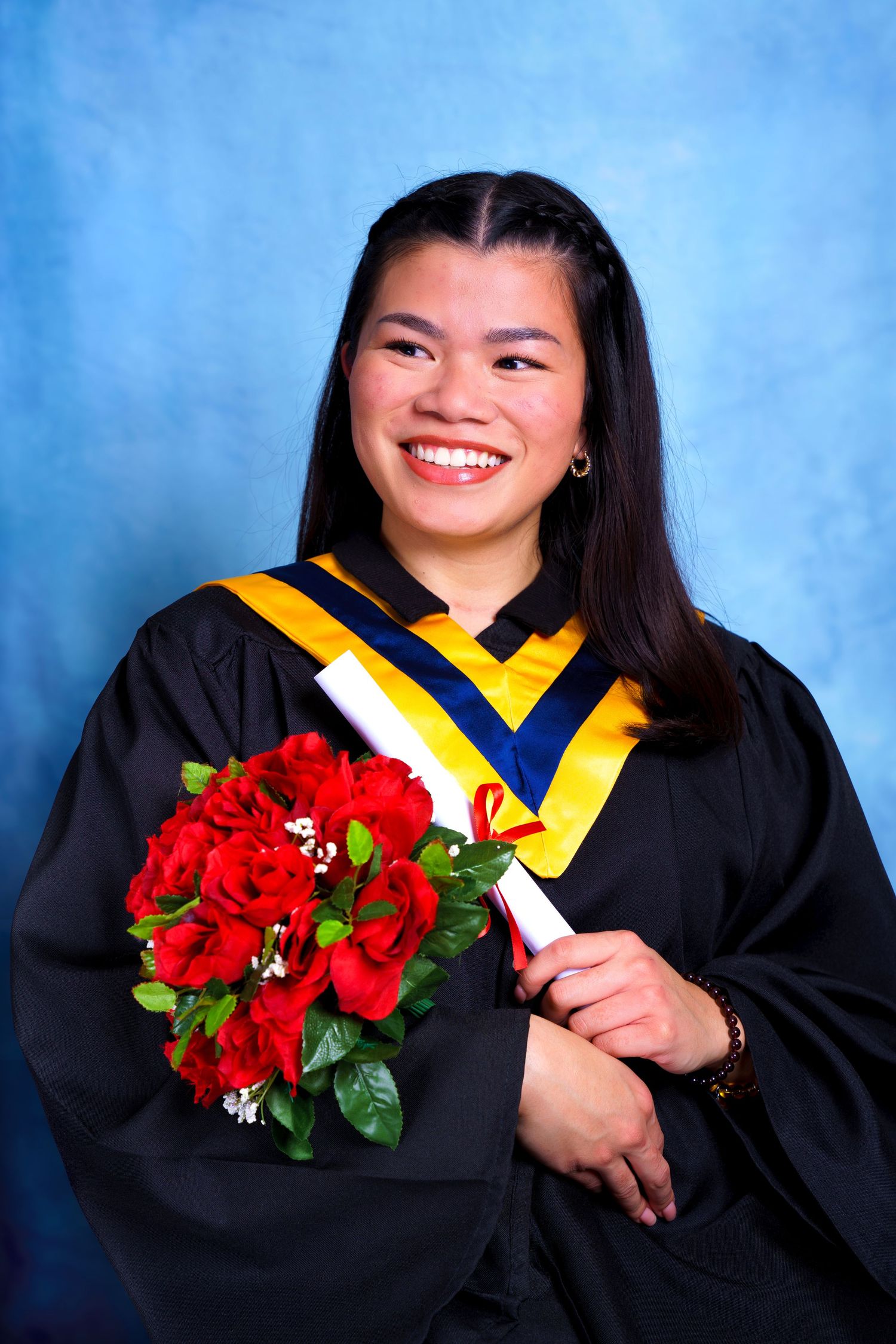 Graduate in black academic regalia holding red bouquet poses against blue background.