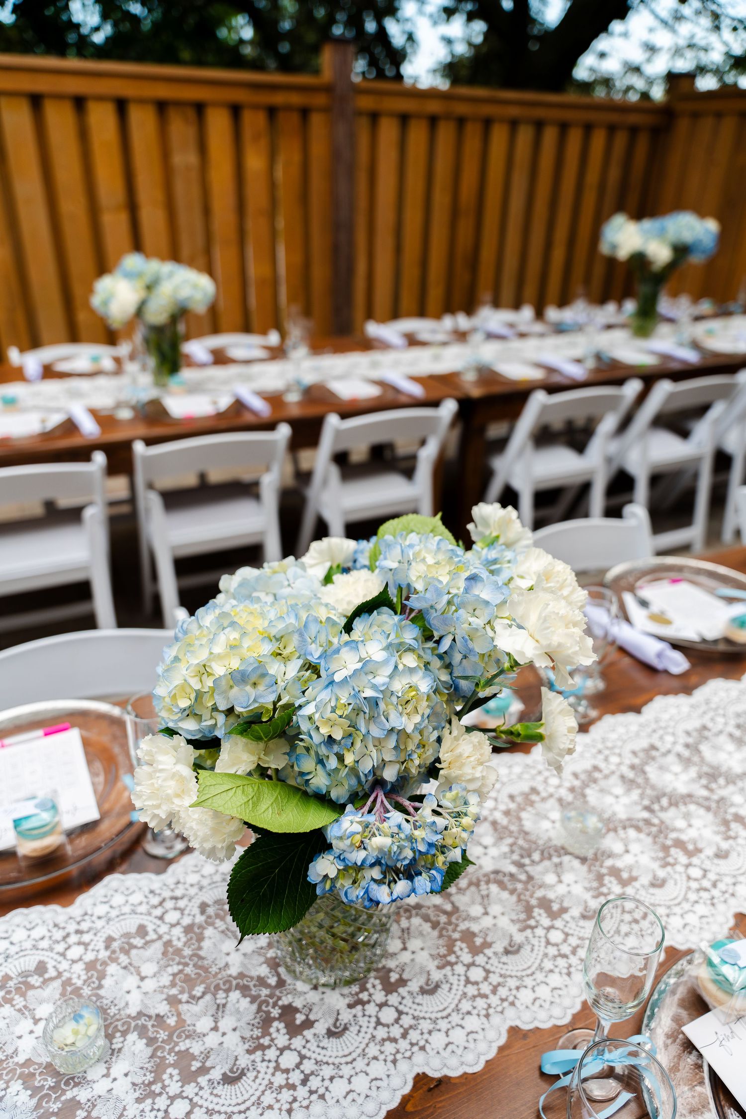 Table settings with blue hydrangea centerpieces and lace runners for an outdoor event.