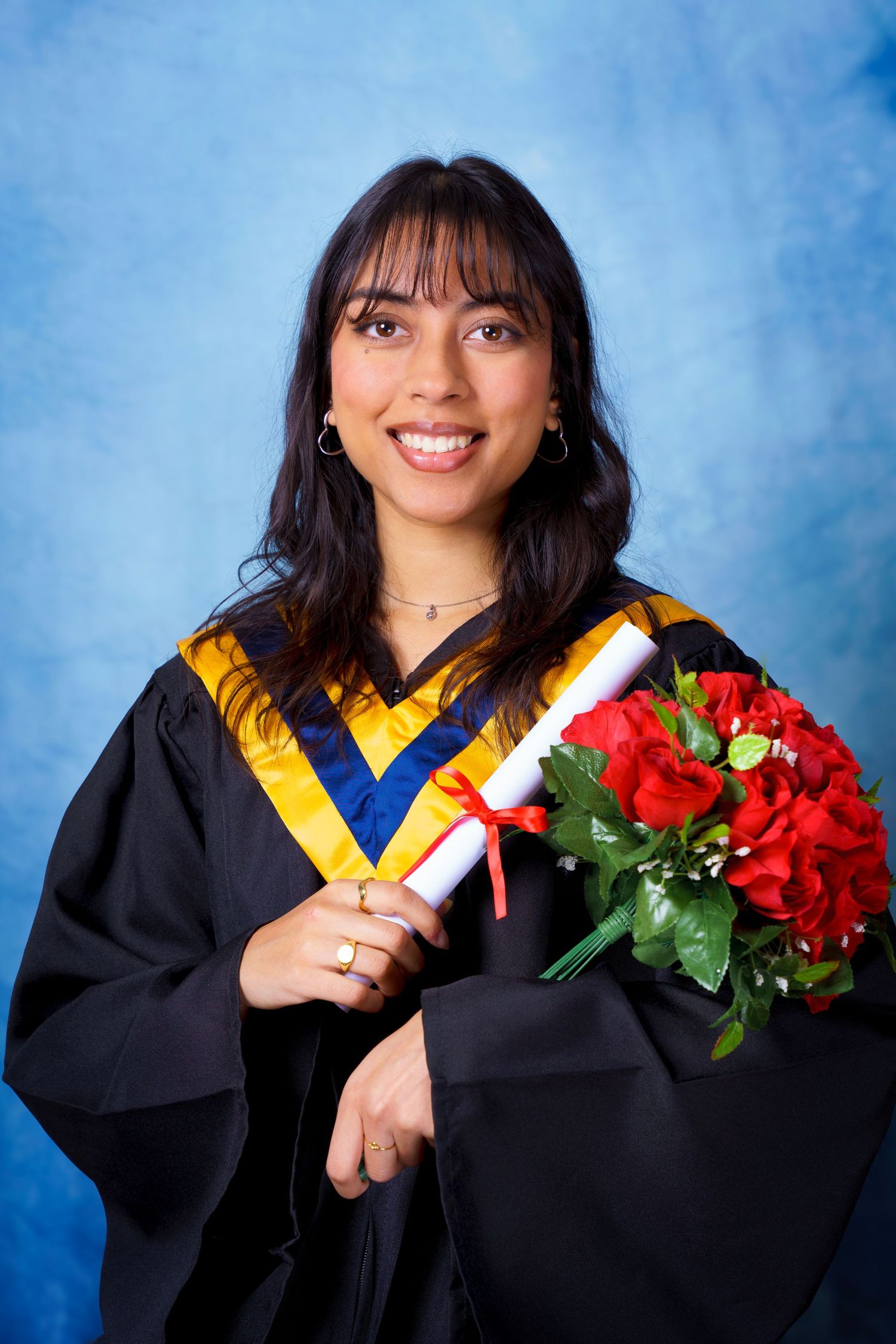 Graduate in black and gold regalia holds diploma and red roses against blue backdrop.