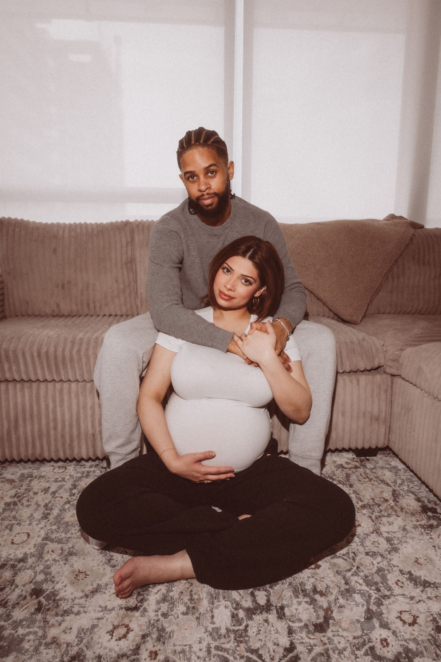 A couple sits together on a grey couch, with the pregnant woman in front wearing black pants and a white top.
