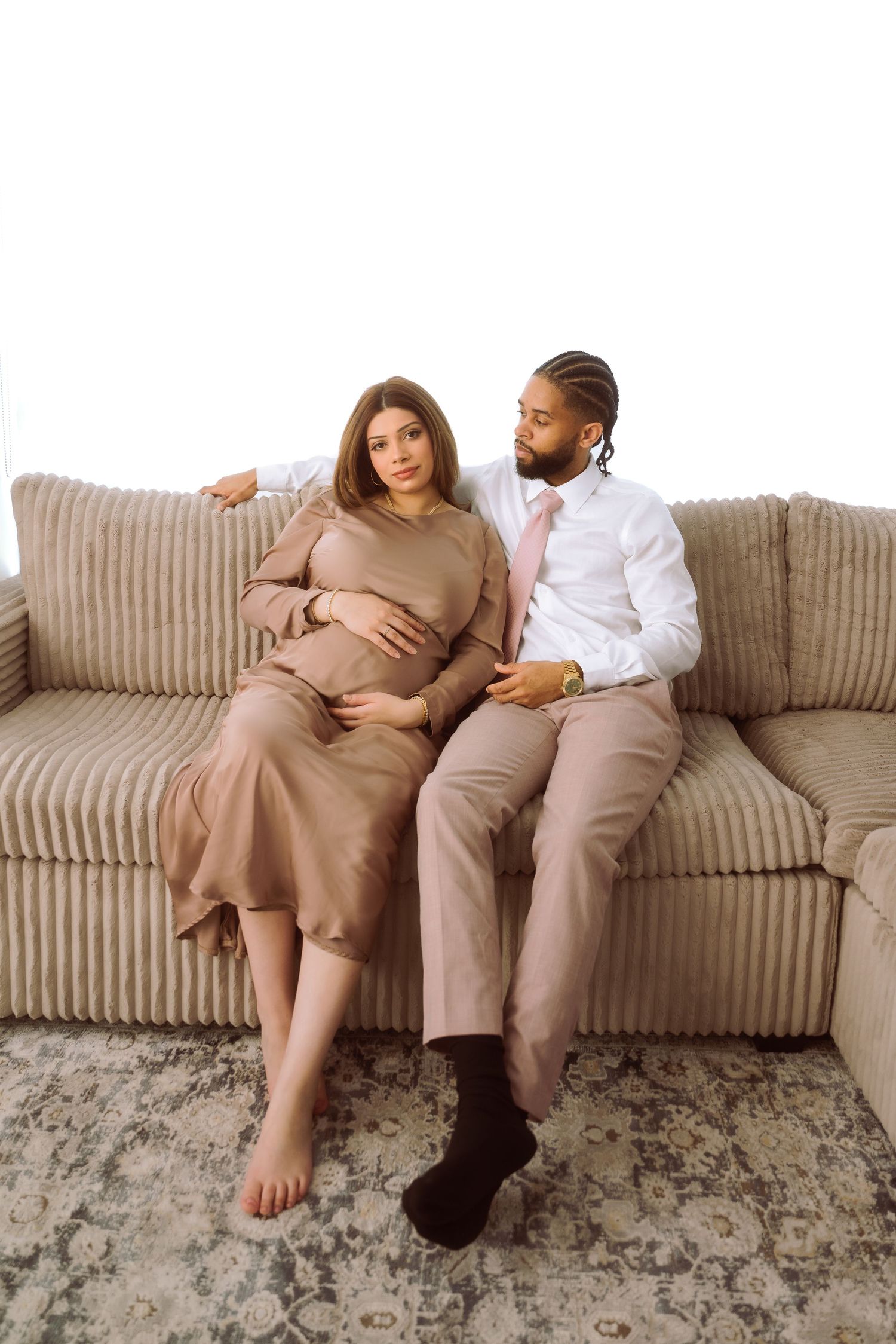 A couple relaxes together on a beige striped couch wearing neutral-toned outfits.