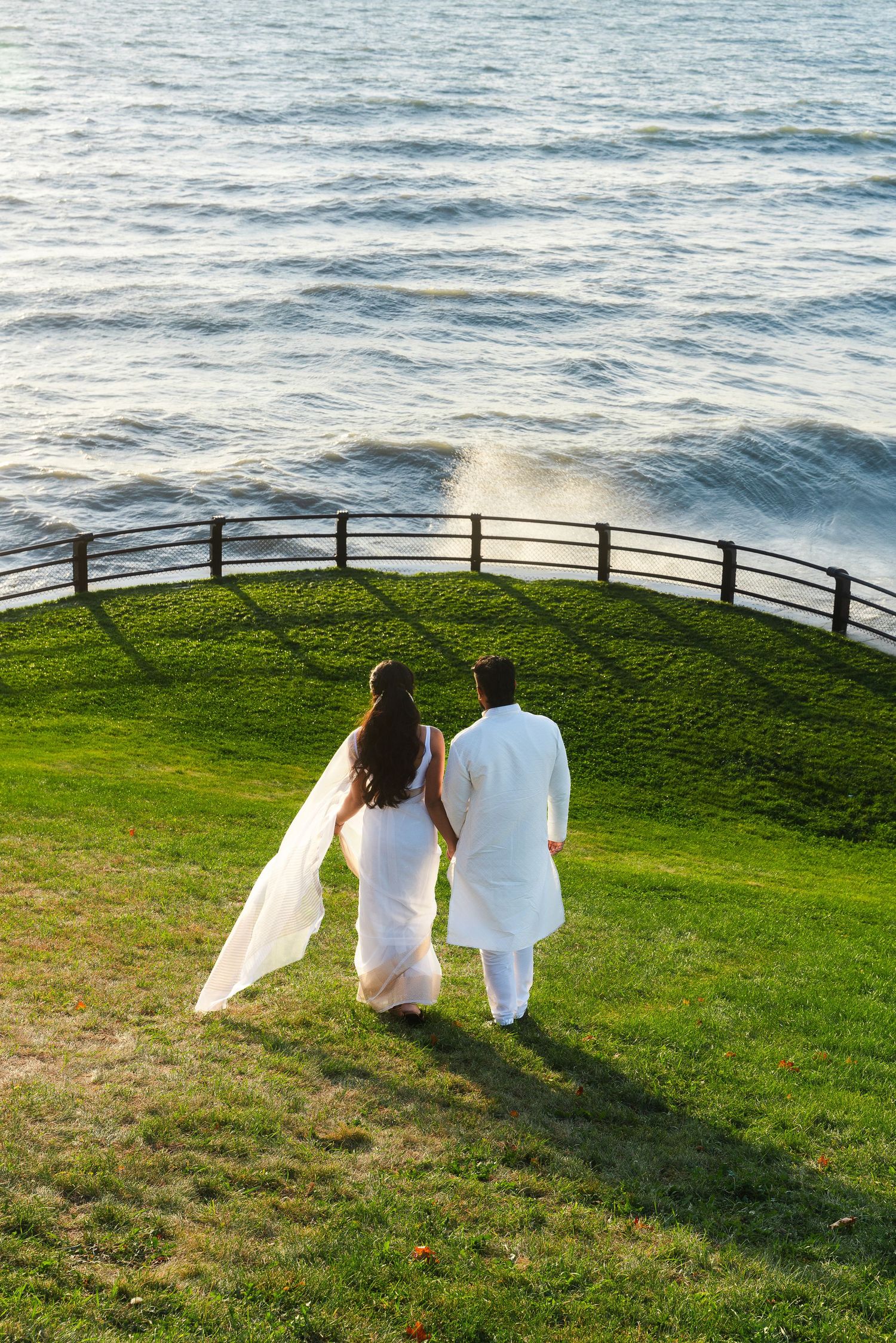 Couple in white walking along grassy oceanfront path with railing.