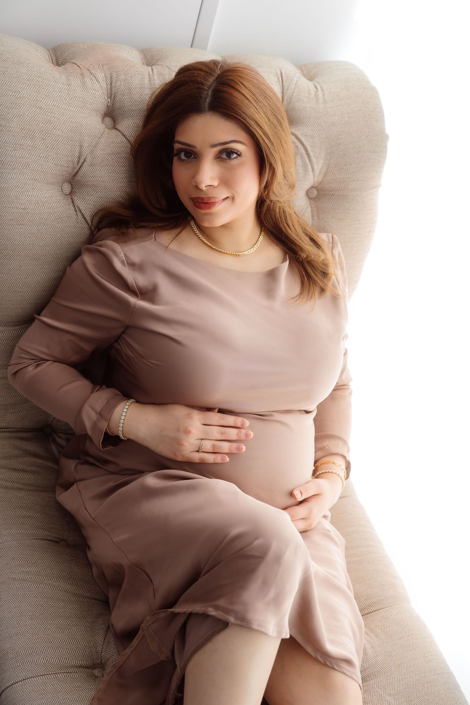 A pregnant woman in beige sweater lounges comfortably on light colored couch in warm, minimalist setting.