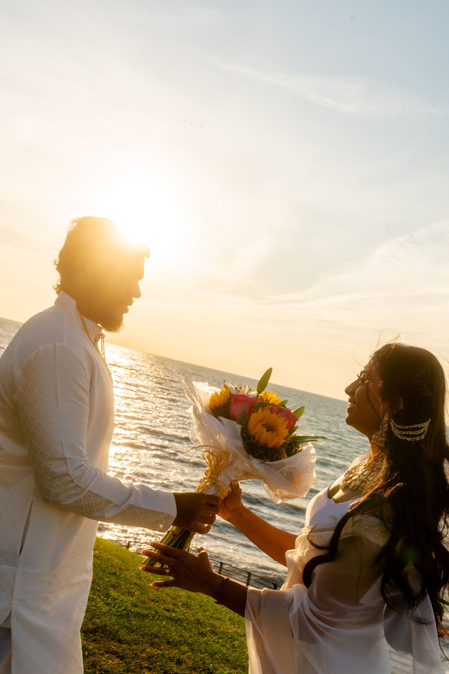 Romantic sunset photoshoot with couple exchanging flowers by the ocean.
