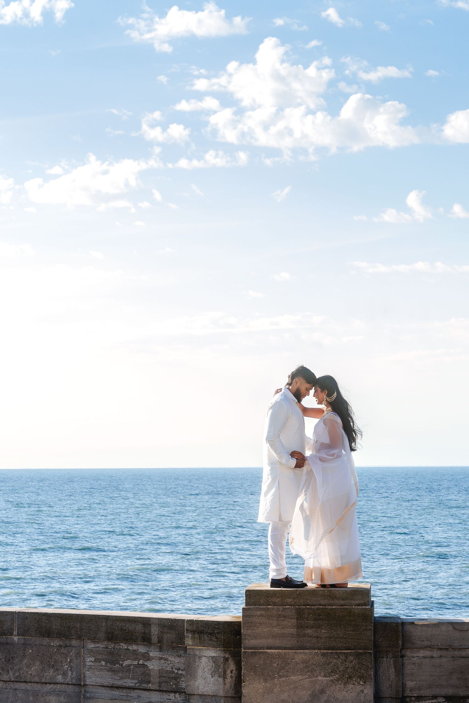 A couple in white attire poses playfully on a stone wall overlooking the ocean.