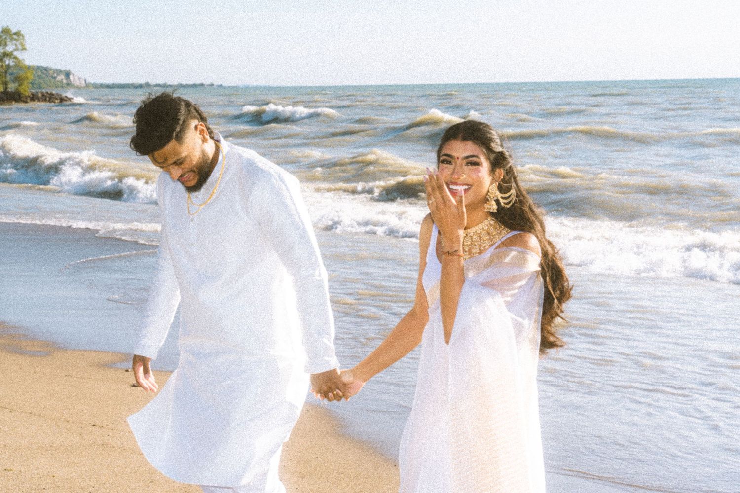 A couple in white outfits runs joyfully along a sunny beach.