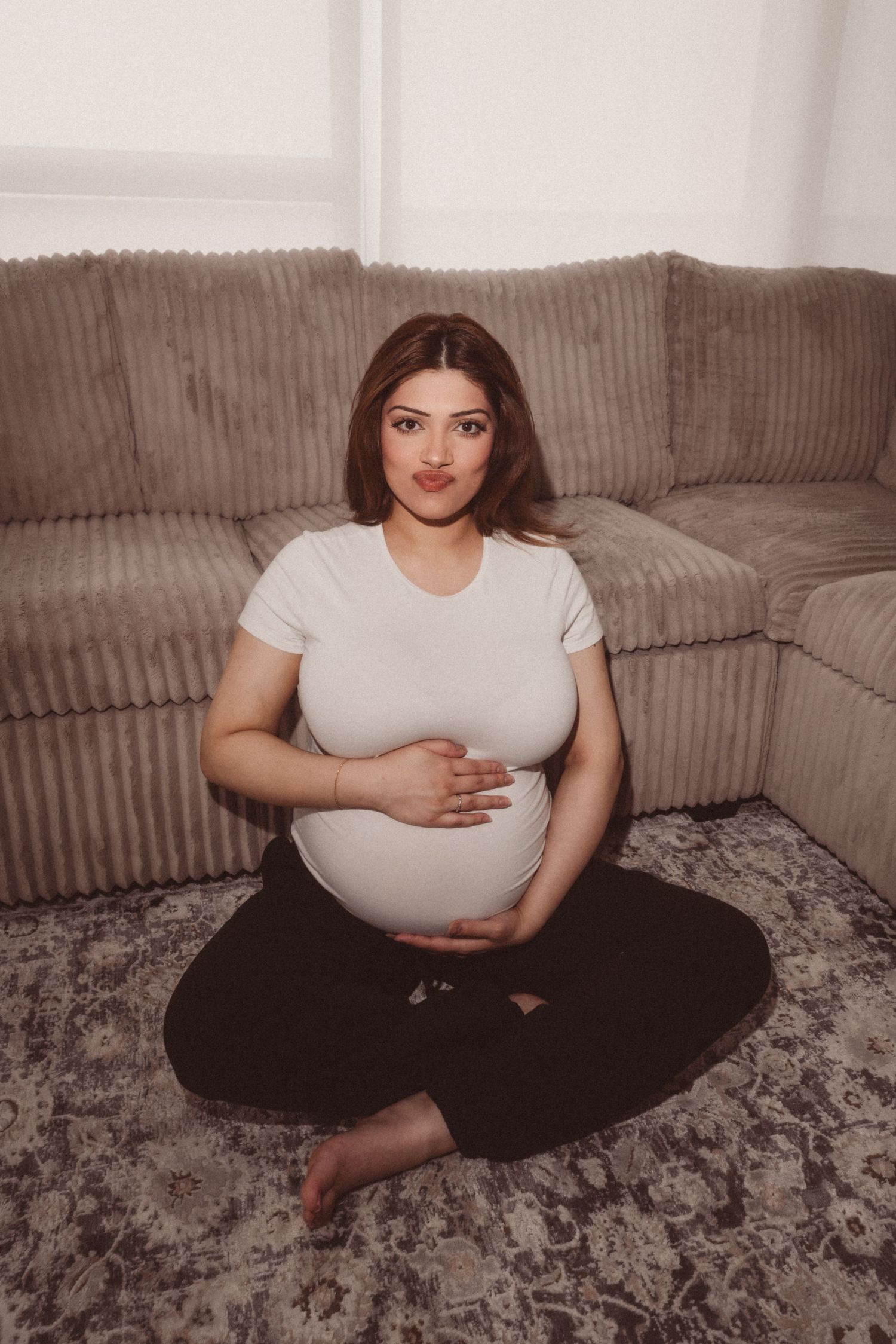 A pregnant woman in a white top and black pants sits cross-legged on a textured gray rug in a home setting.
