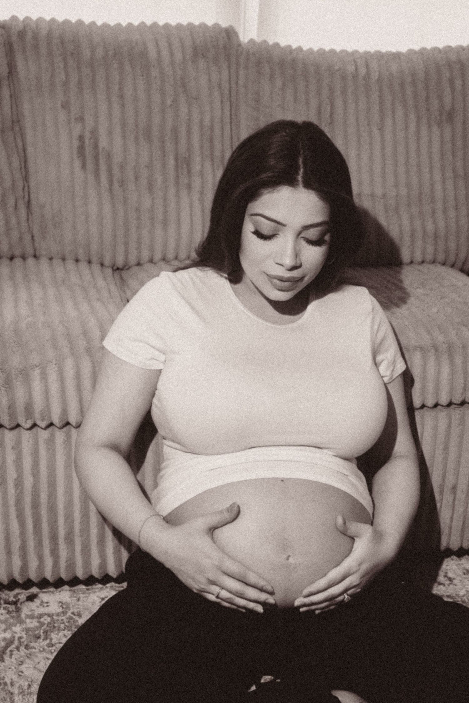 A black and white photo shows an expectant mother in a white t-shirt sitting on a couch and cradling her pregnant belly.