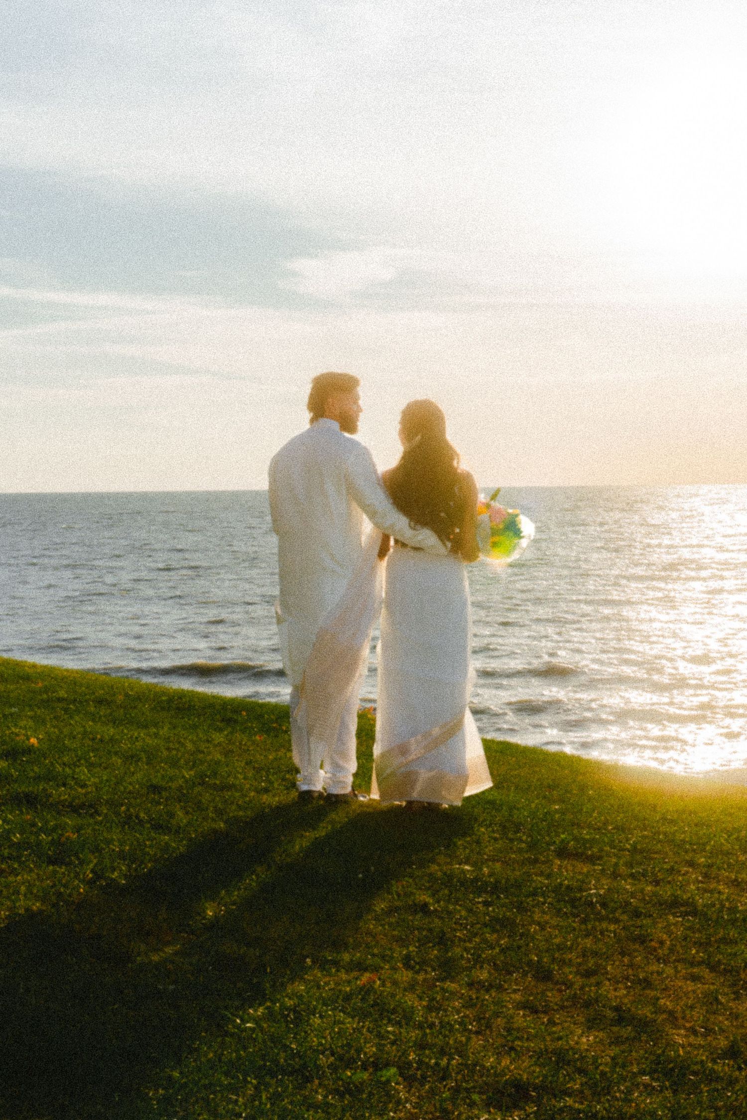 Couple in white attire sitting together on oceanside cliff during golden hour.