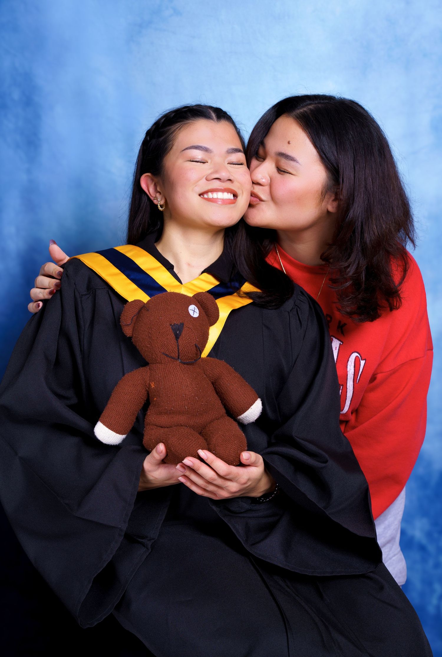Two people sharing a joyful moment at graduation while holding a brown teddy bear in academic regalia against blue backdrop.