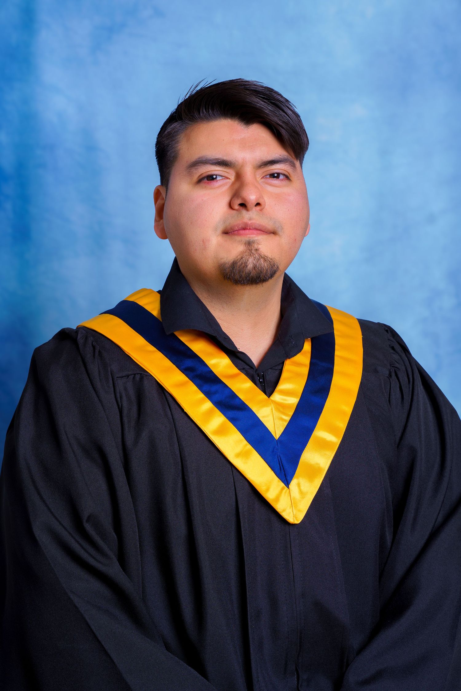 Graduate in black academic gown with gold and blue chevrons poses formally against blue backdrop.
